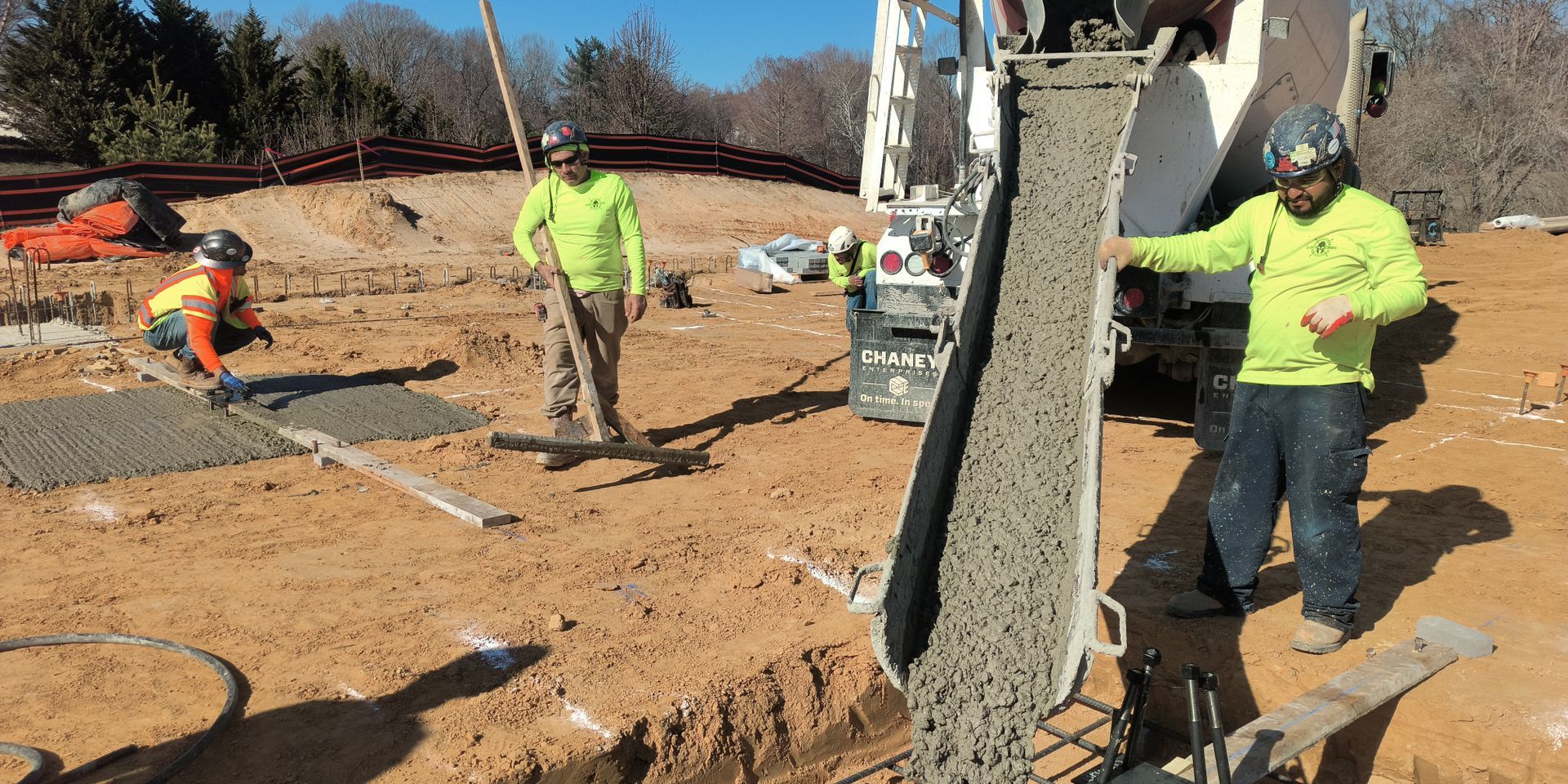 Construction workers pouring concrete at a roadside site, with one worker guiding the mix from a chute.