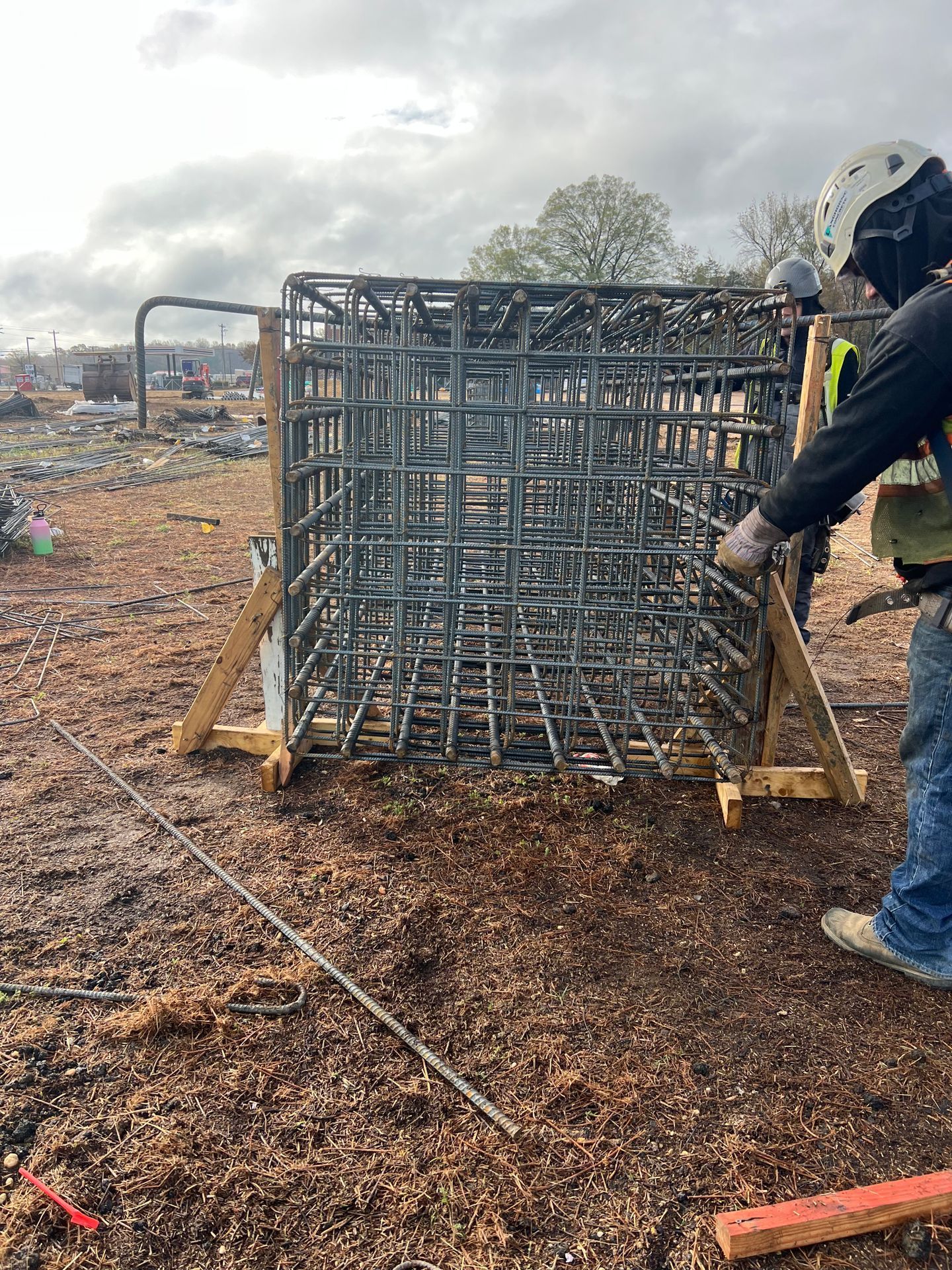 Worker moving a large metal cage frame on a muddy construction site under cloudy skies