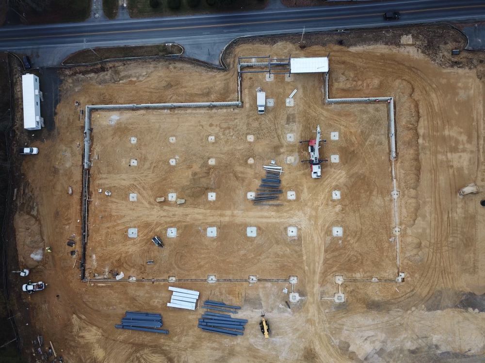 Aerial view of a construction site with foundation markers, excavator, and dirt lots near a road.