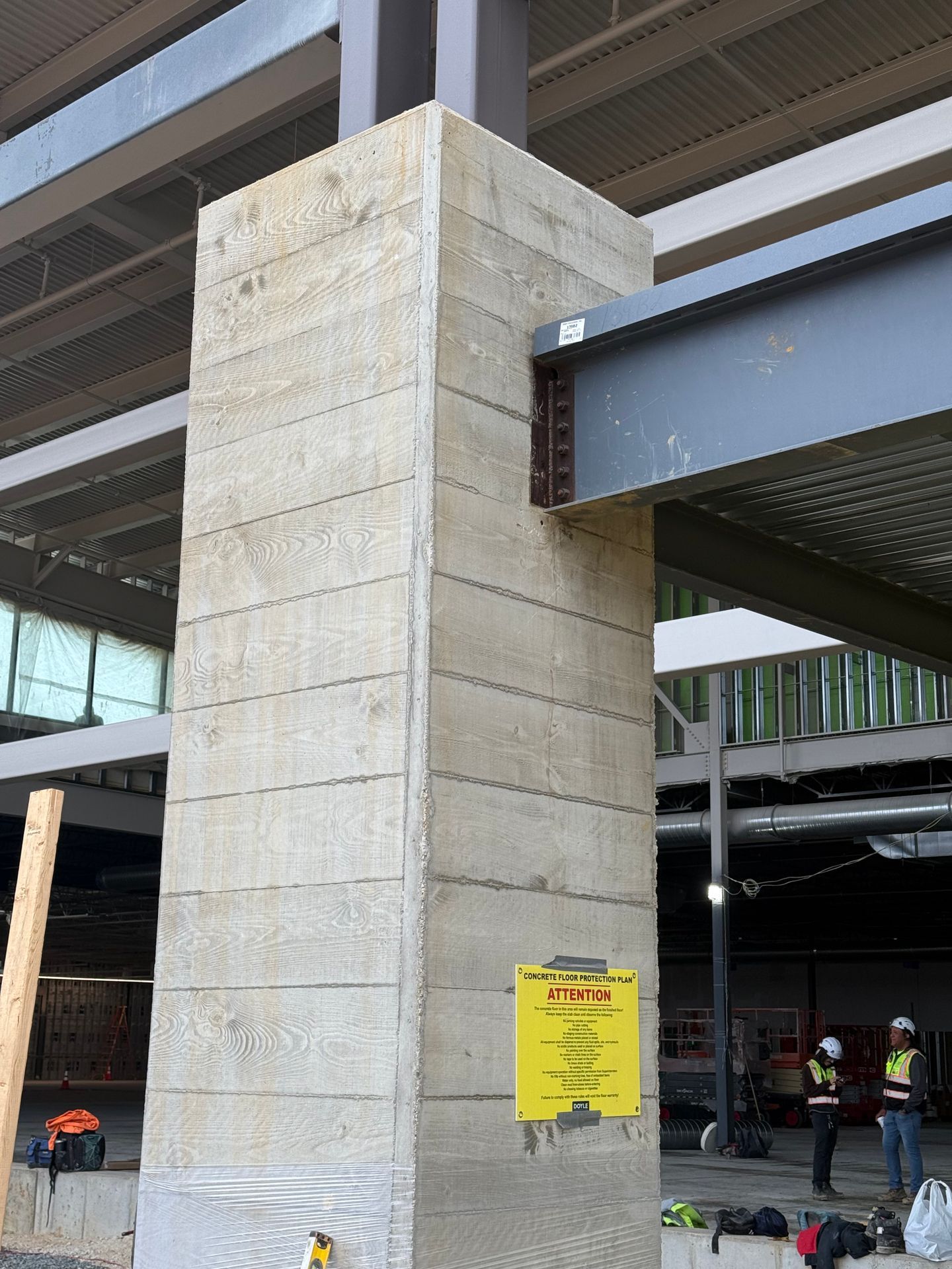Concrete pillar with a yellow sign at a construction site, with steel beams and workers in the background