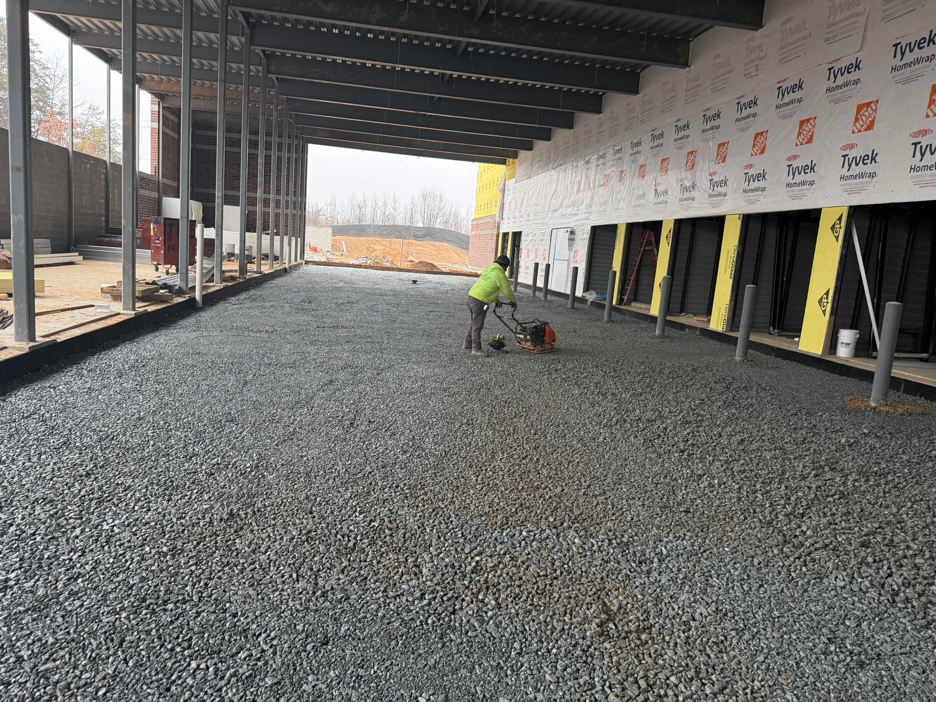 Worker spreading gravel on a construction floor inside a framed building shell