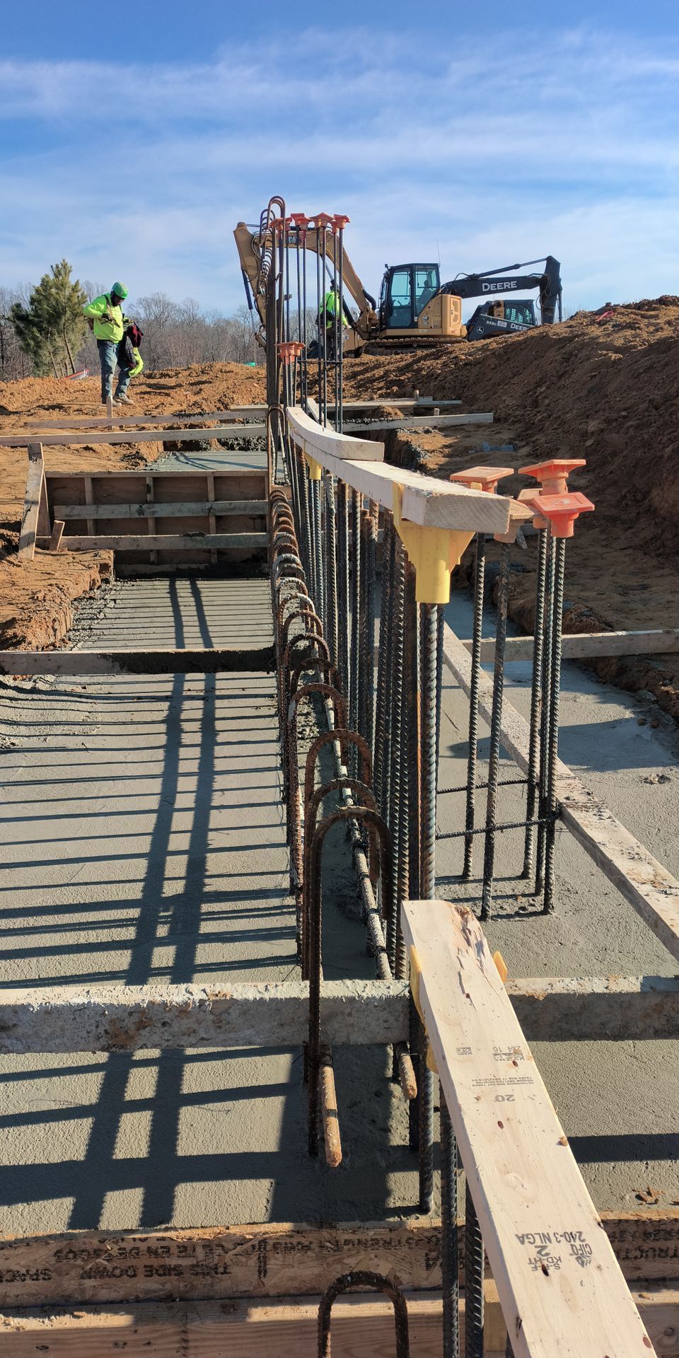 Construction site with concrete foundation formwork, rebar, workers, and excavator on a dirt embankment