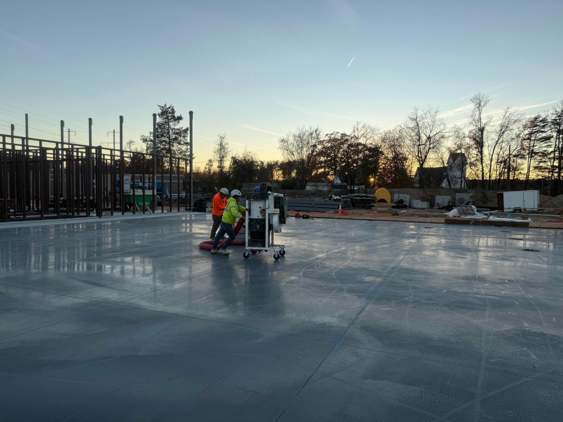 Workers operating equipment on a smooth concrete slab at a construction site at dusk.