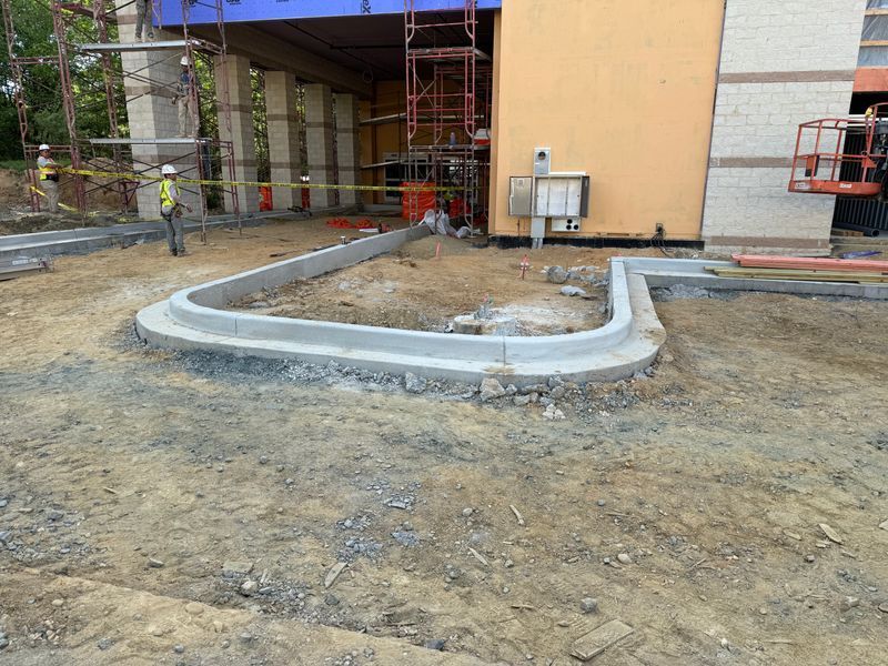 Construction site with curved concrete curb around a dirt patch beside a building and scaffolding