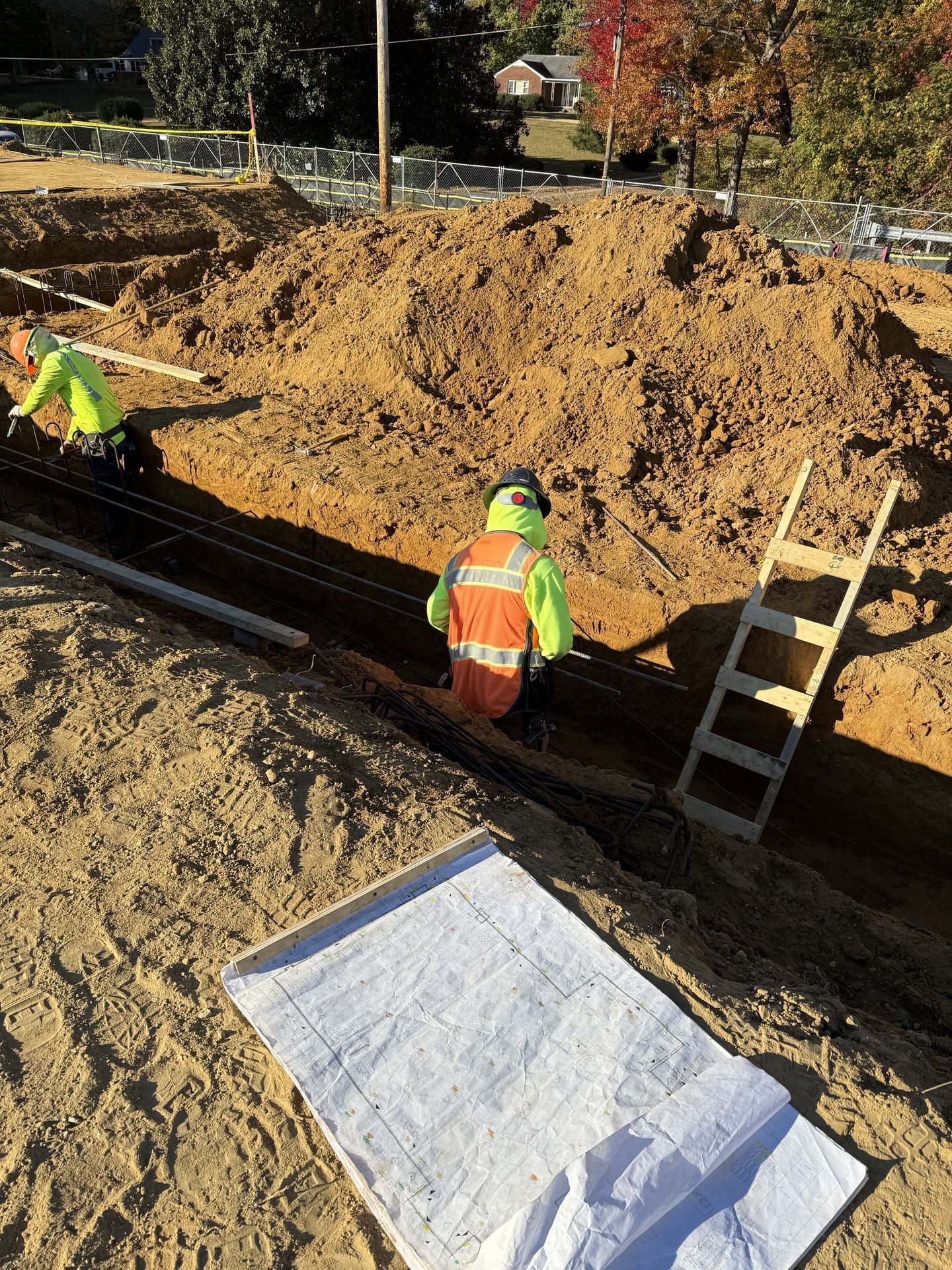 Workers in neon safety vests digging a trench at a construction site with a dirt mound and ladder