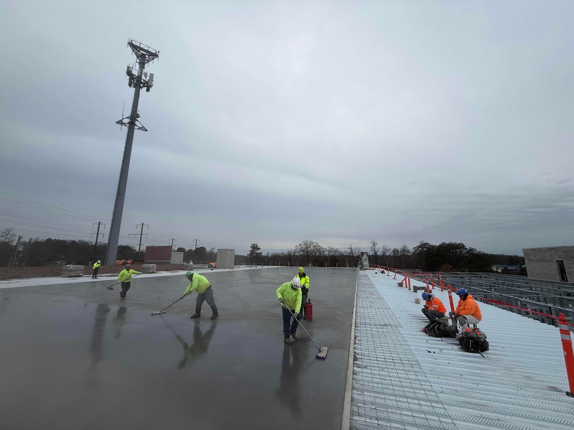Workers in neon vests on a wet rooftop, cleaning near orange safety barriers under a cloudy sky