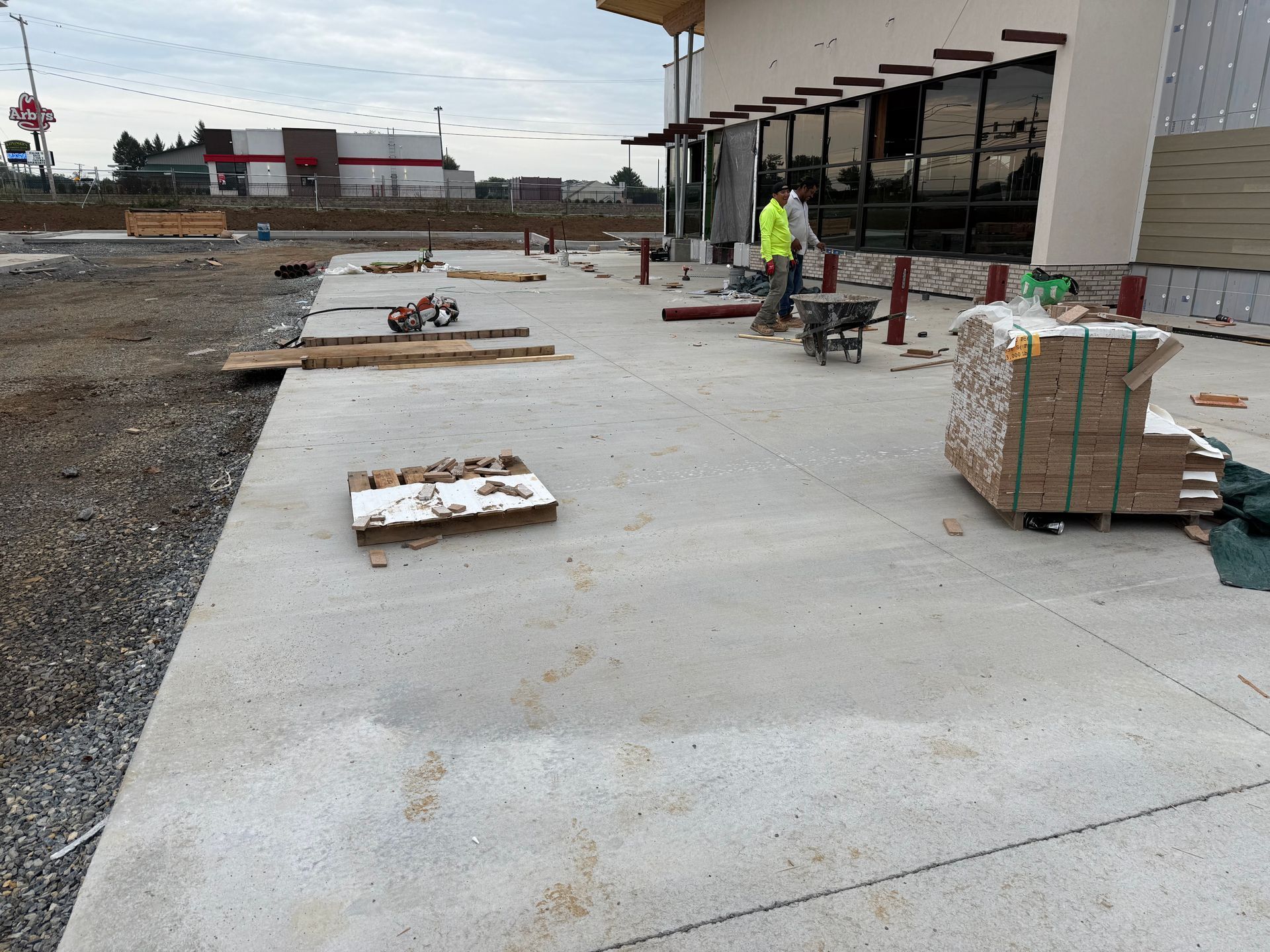 New storefront sidewalk under construction with workers, pallets, and building materials on a concrete slab