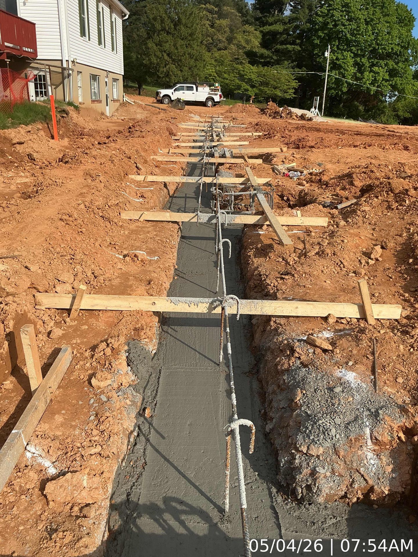 Construction trench with rebar and wooden supports beside a house on a dirt lot