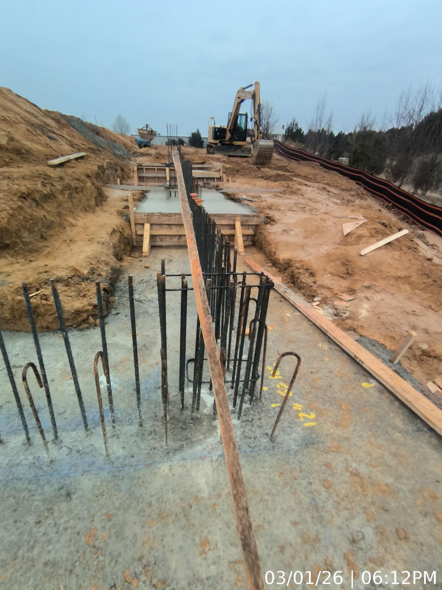 Construction site with rebar and concrete slab in foreground, trench and excavator in the distance