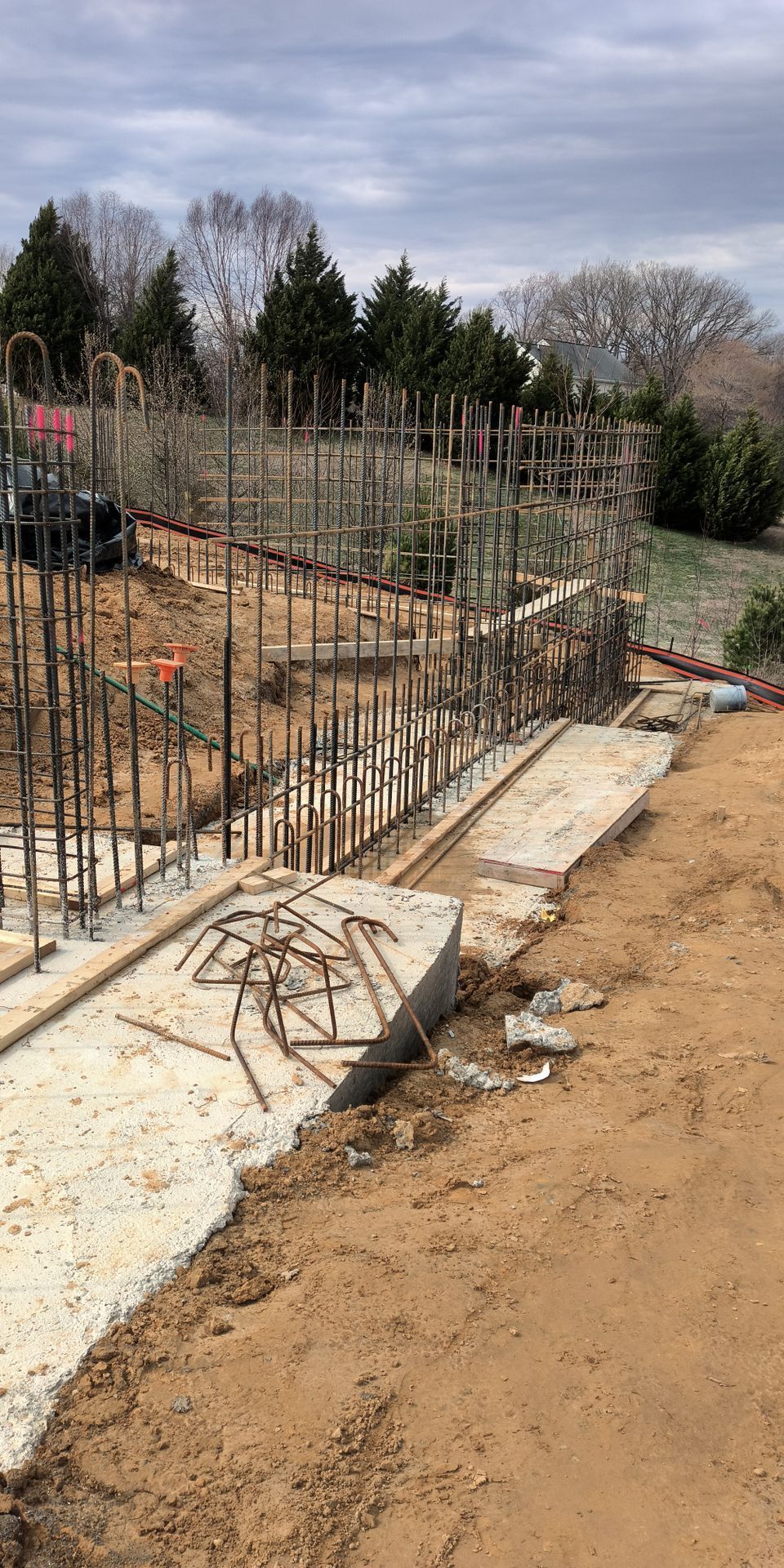 Construction site with steel rebar columns and concrete footing beside a dirt path and trees
