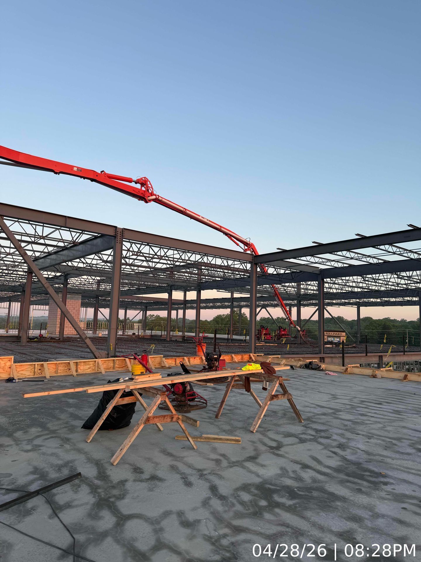 Construction site with steel frame, red concrete pump boom, and sawhorses on a concrete slab at sunset