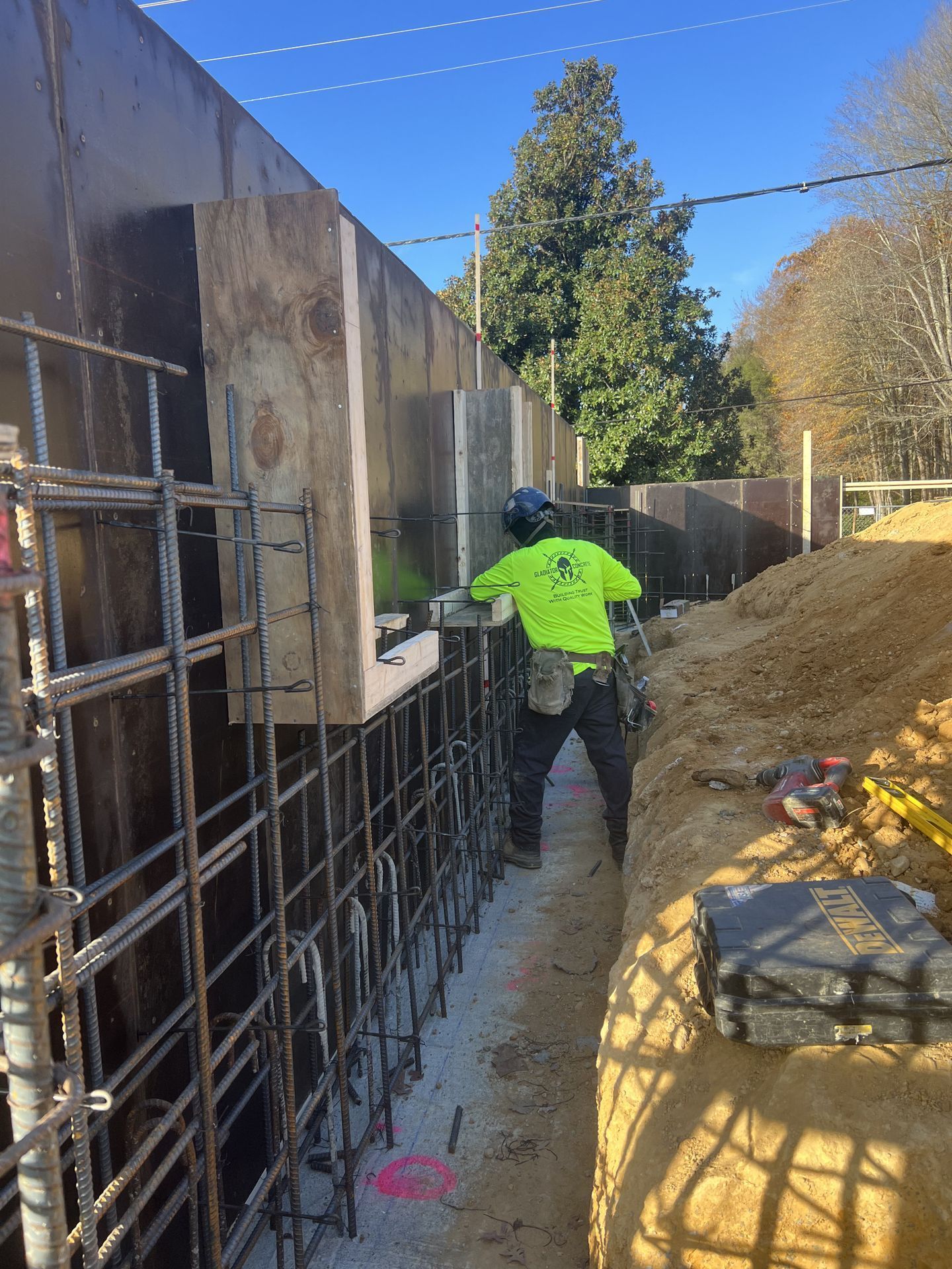 Construction worker in a neon jacket on a narrow concrete walkway beside rebar and a retaining wall
