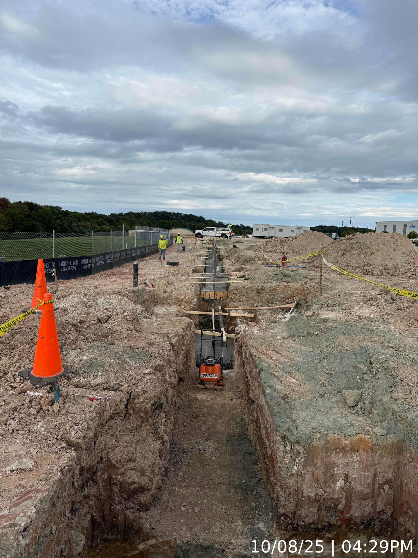 Construction trench with orange cone, excavator, and survey stakes beside a fenced path under cloudy sky