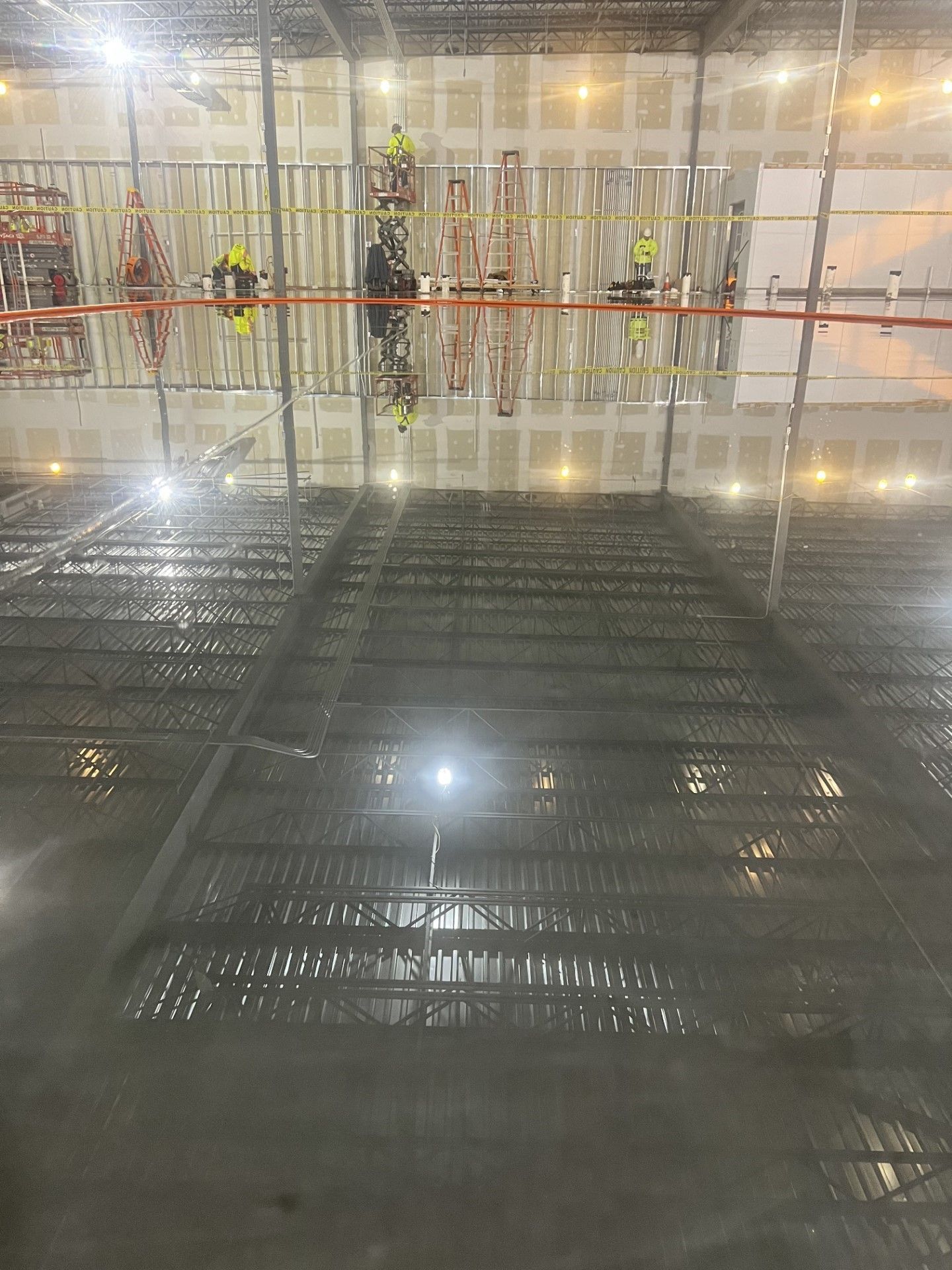 Workers in safety vests on a scaffold inside a construction site with metal framing and wet floors
