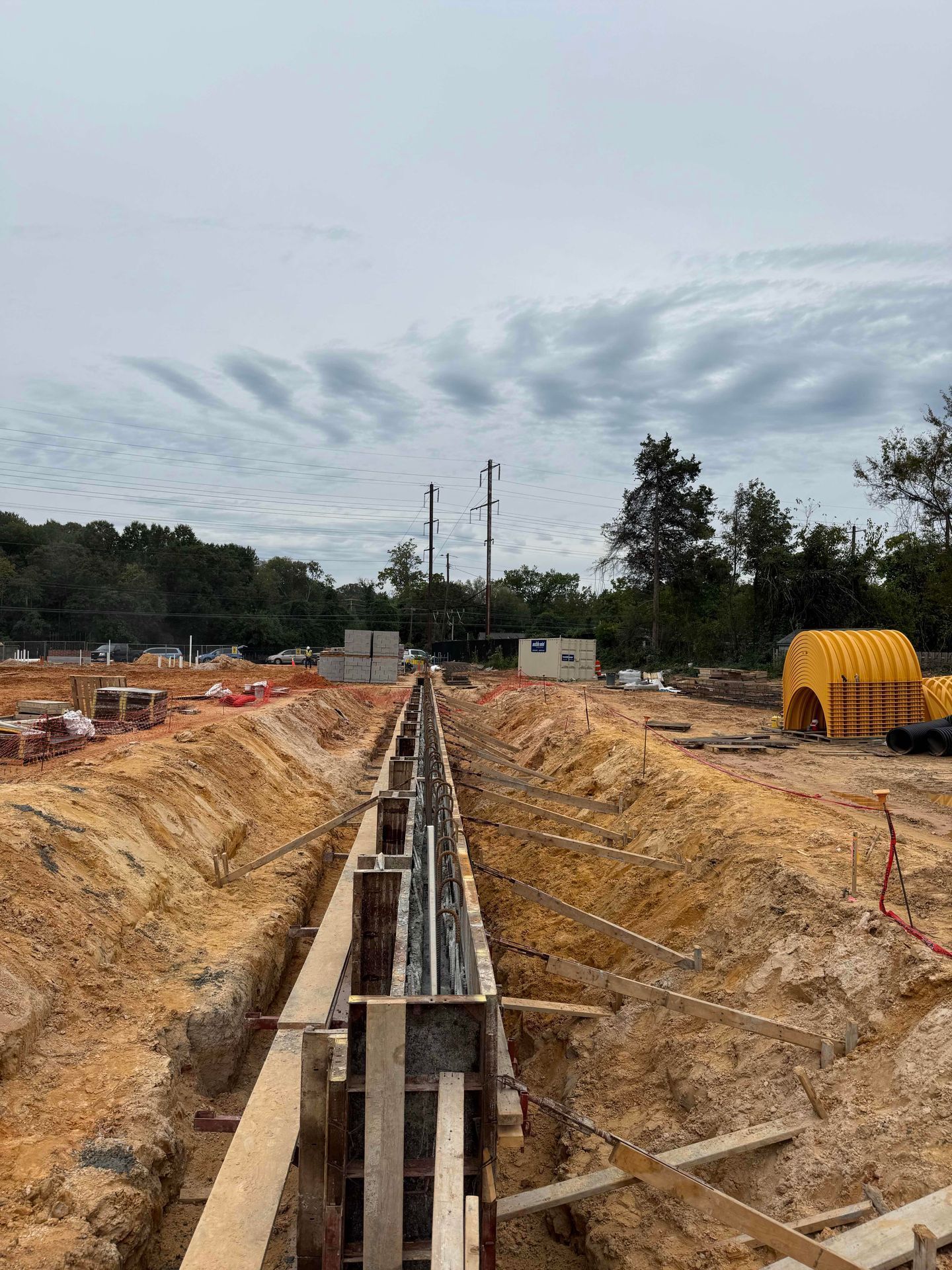 Construction trench with a long concrete formwork channel, dirt walls, and equipment at a worksite under a cloudy sky