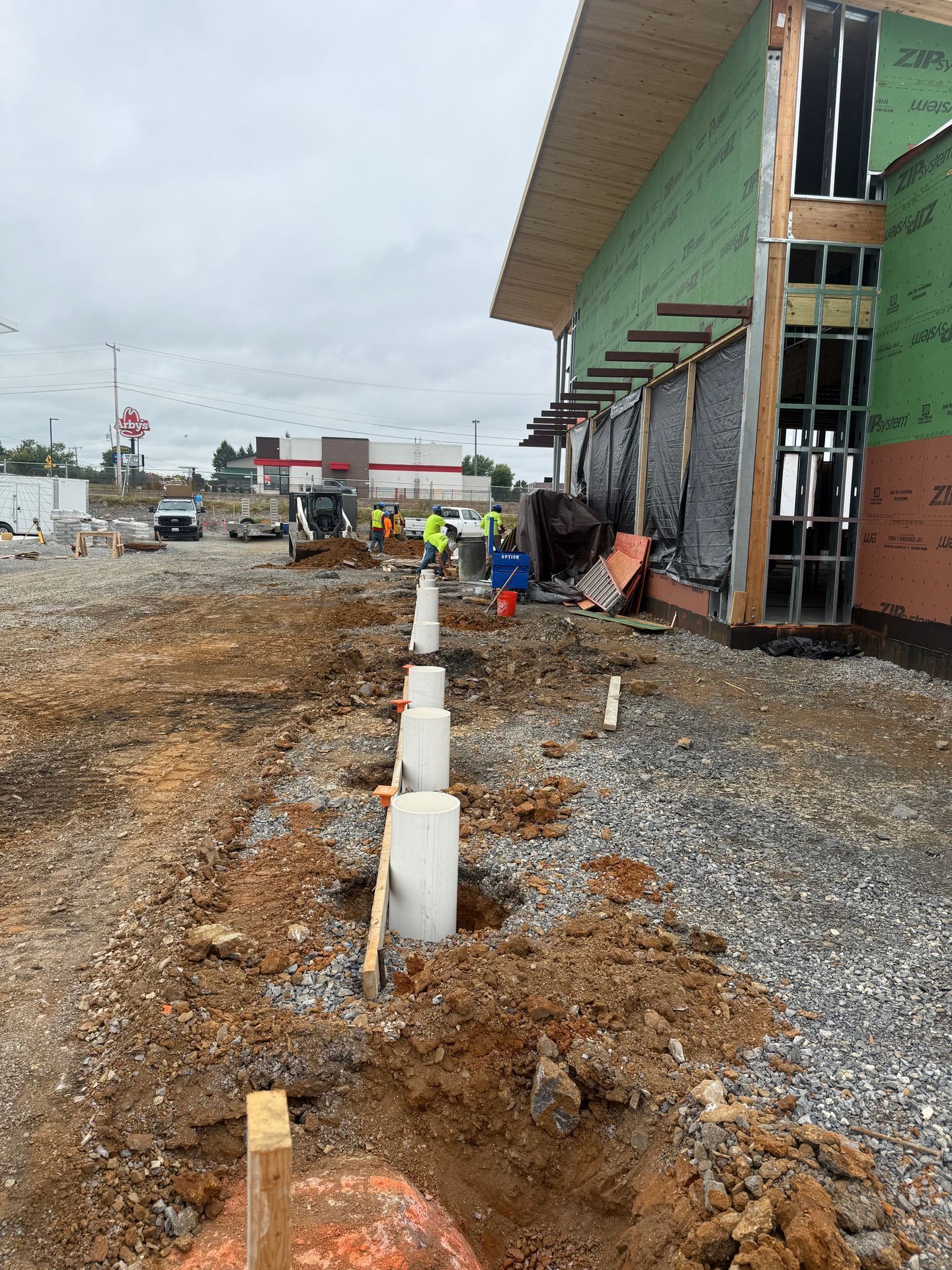 Construction site beside green building with workers, dirt path, and white pipes along the ground