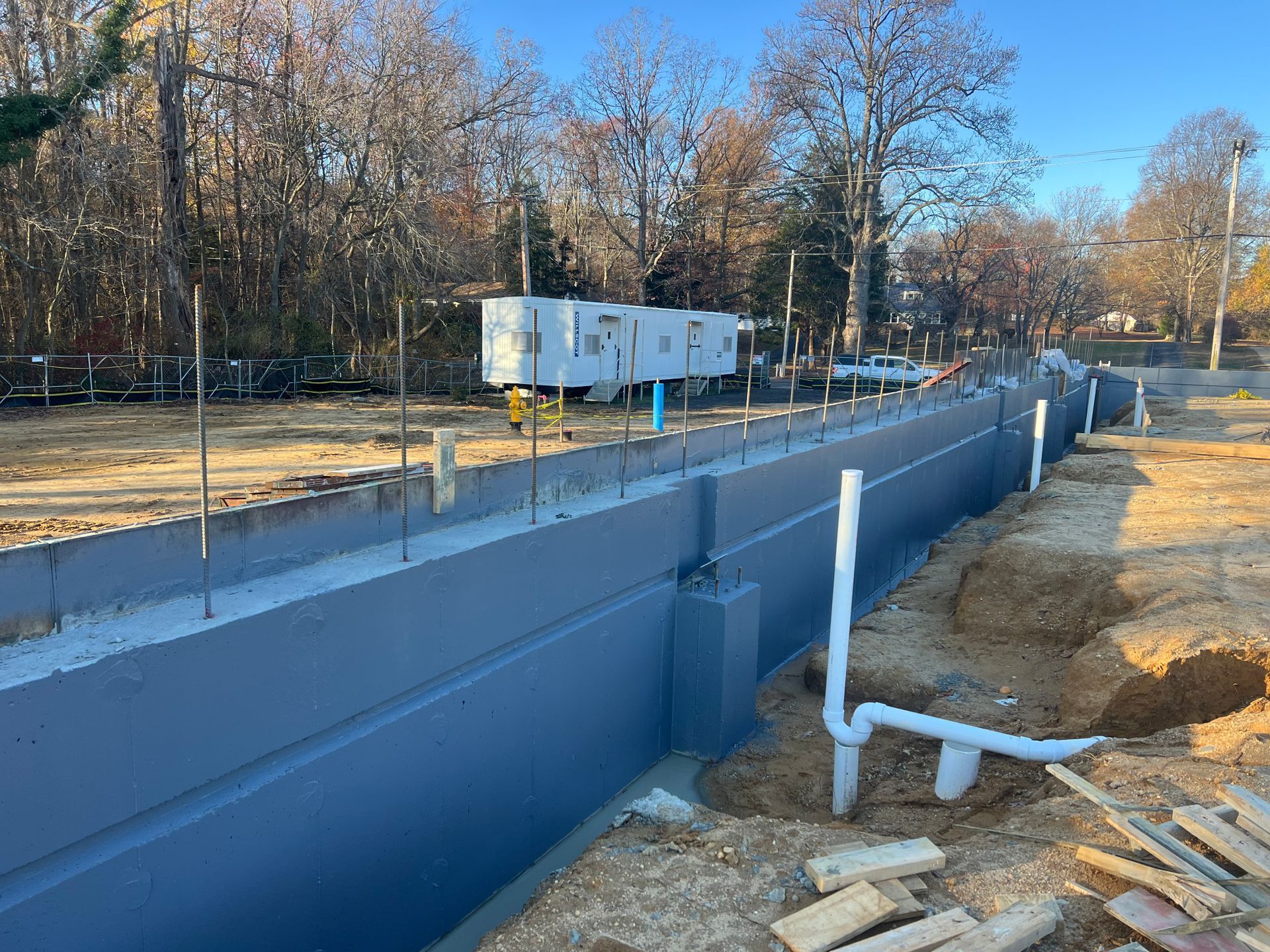 Blue concrete foundation walls at a construction site, with dirt, piping, and a white trailer in the background