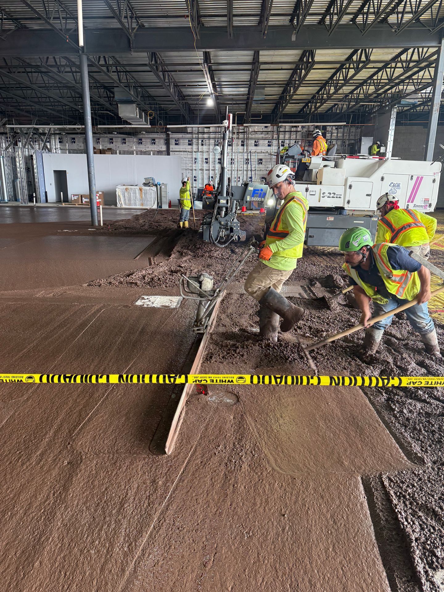 Workers in neon vests spread wet concrete in a large indoor construction site.