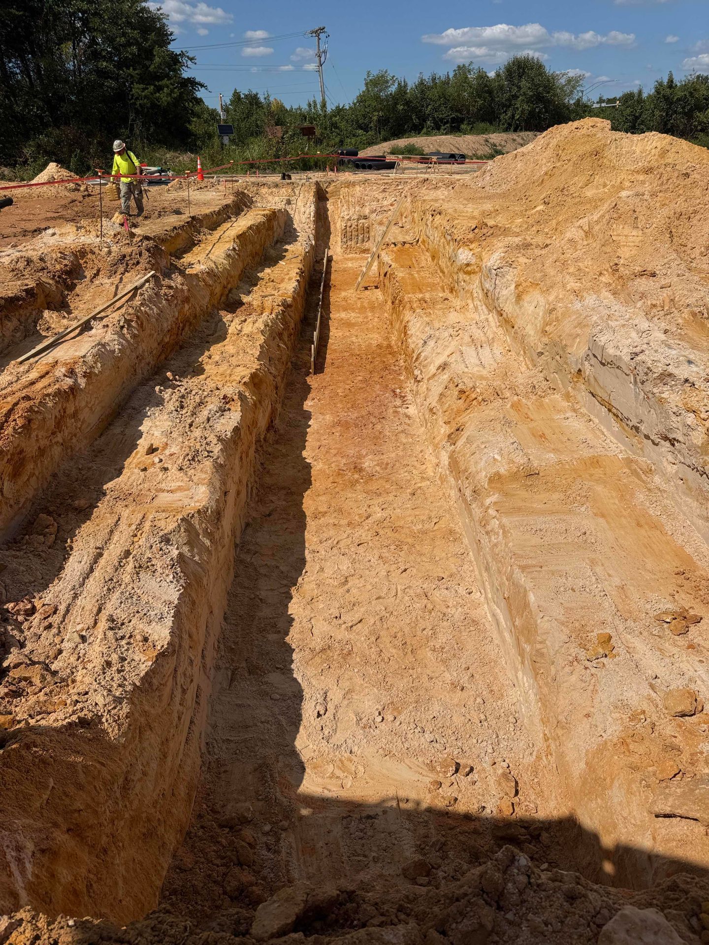 Deep trench excavation in sandy soil with a worker in a yellow vest under a blue sky.