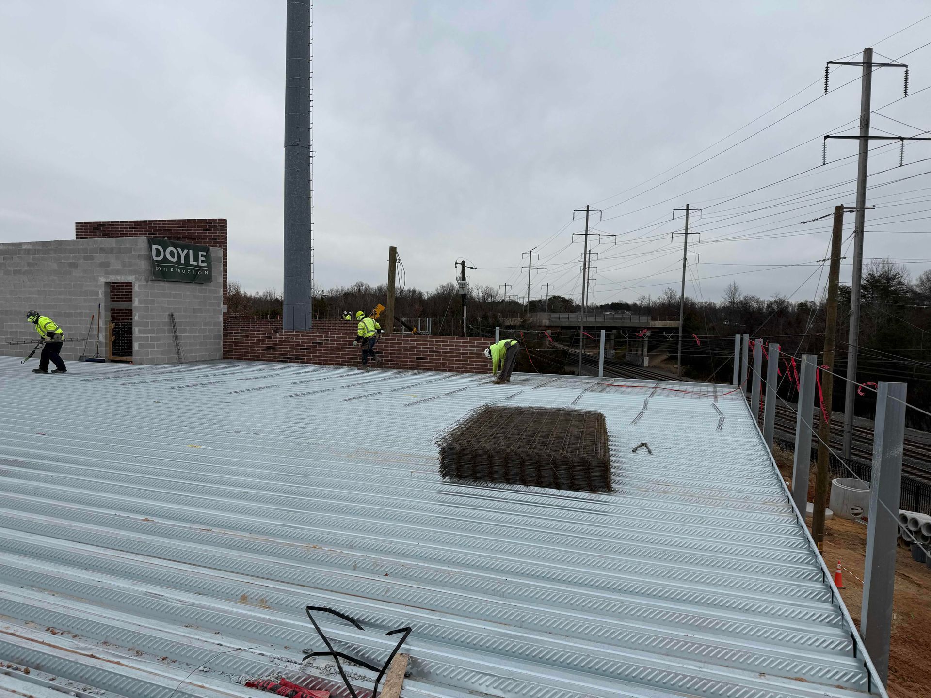 Workers on a corrugated metal roof near a brick building and utility poles on an overcast day