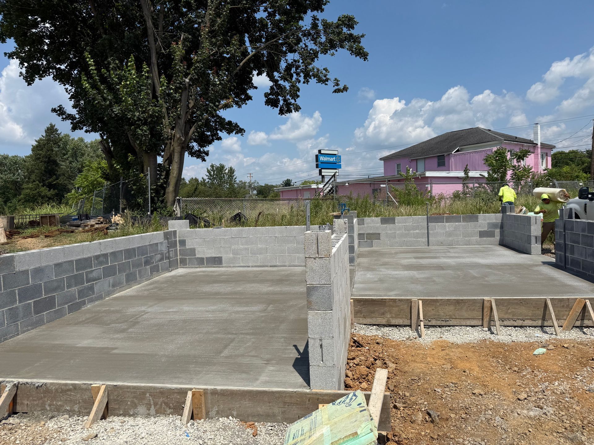Concrete foundation under construction with cinder block walls, a large tree, and a house in the background