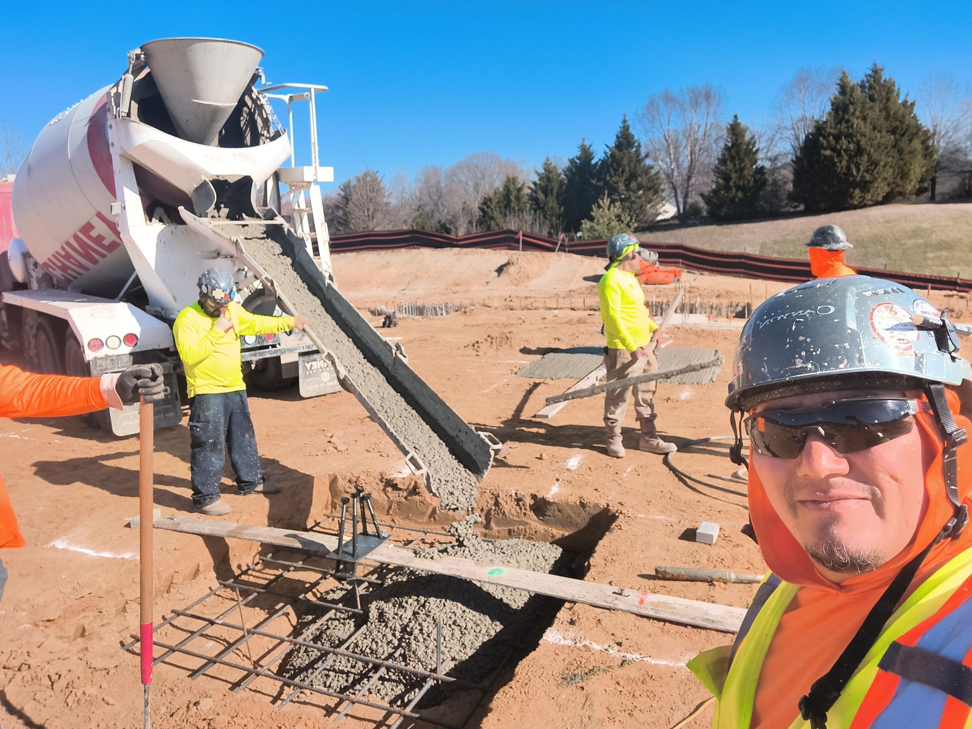 Construction workers guiding concrete from a mixer truck at a site, with one worker taking a selfie in the foreground