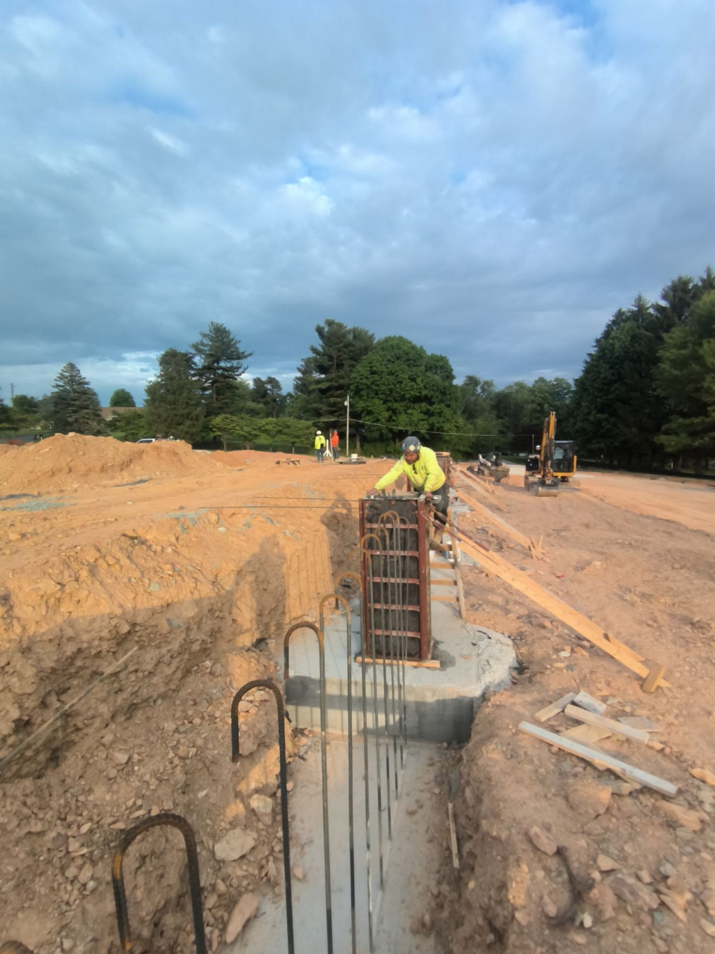 Construction site with workers near exposed concrete rebar and dirt trench under a cloudy sky