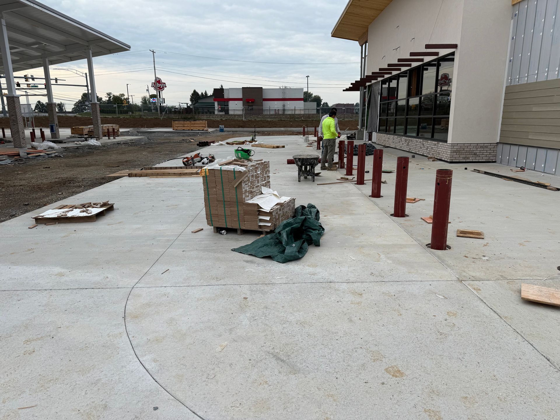 Construction site with concrete walkway, red bollards, stacked bricks, and workers in the background