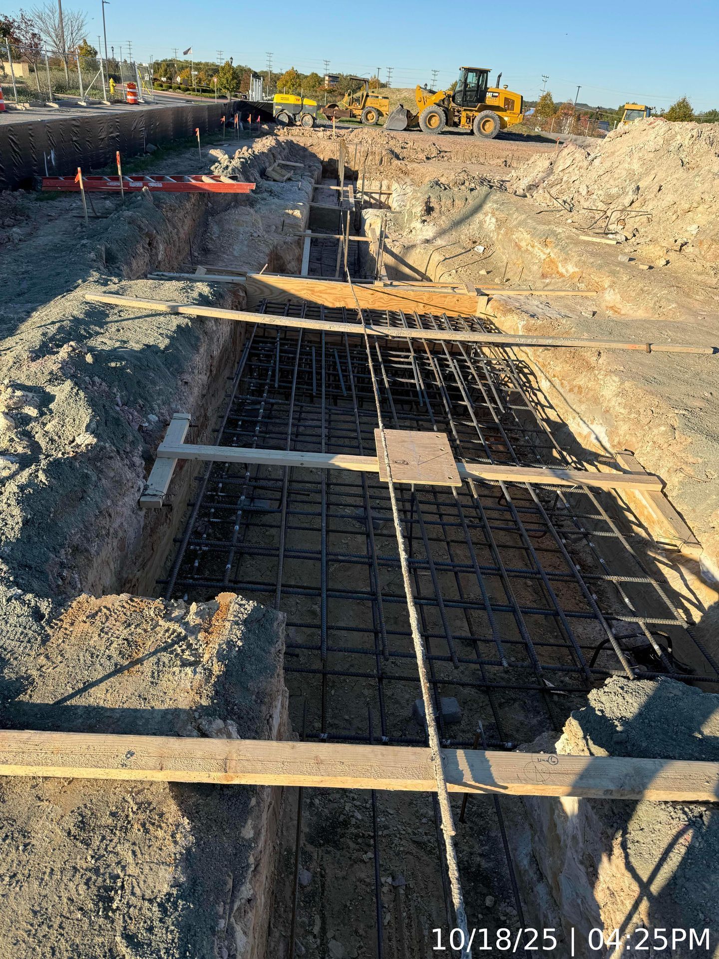 Construction trench with rebar and wooden supports beside a dirt roadway under excavation.