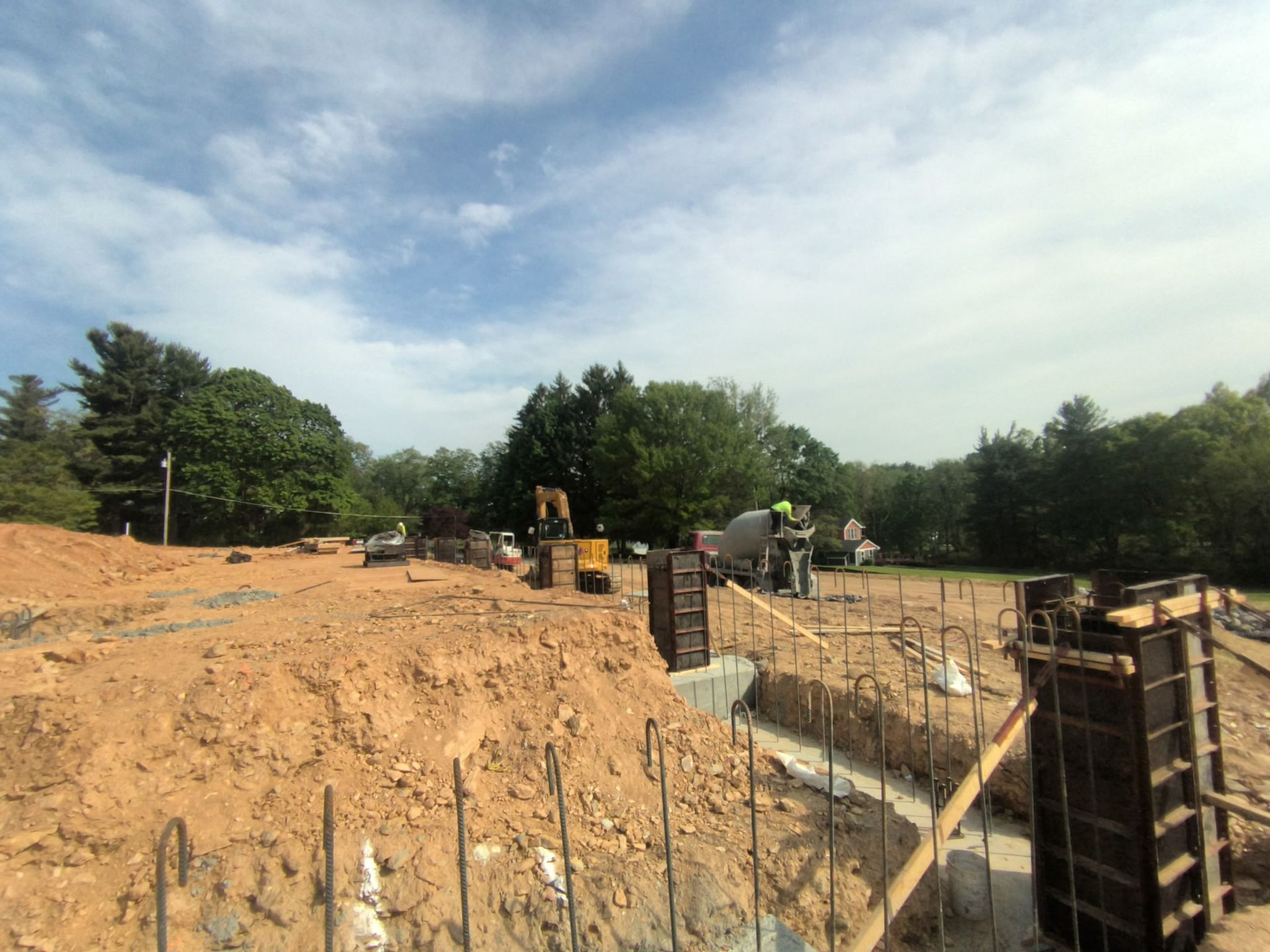 Construction site with excavators, rebar, and wooden forms beside a dirt excavation under a cloudy sky