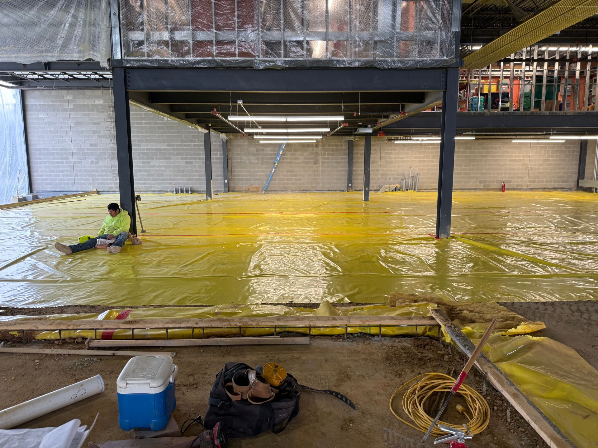 Indoor construction site with yellow waterproofed floor, metal beams, and workers near tools in the foreground