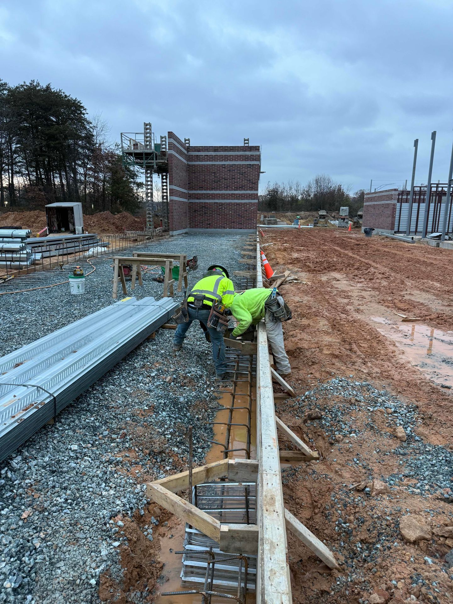 Workers in neon vests install rebar in a foundation trench at a muddy construction site.
