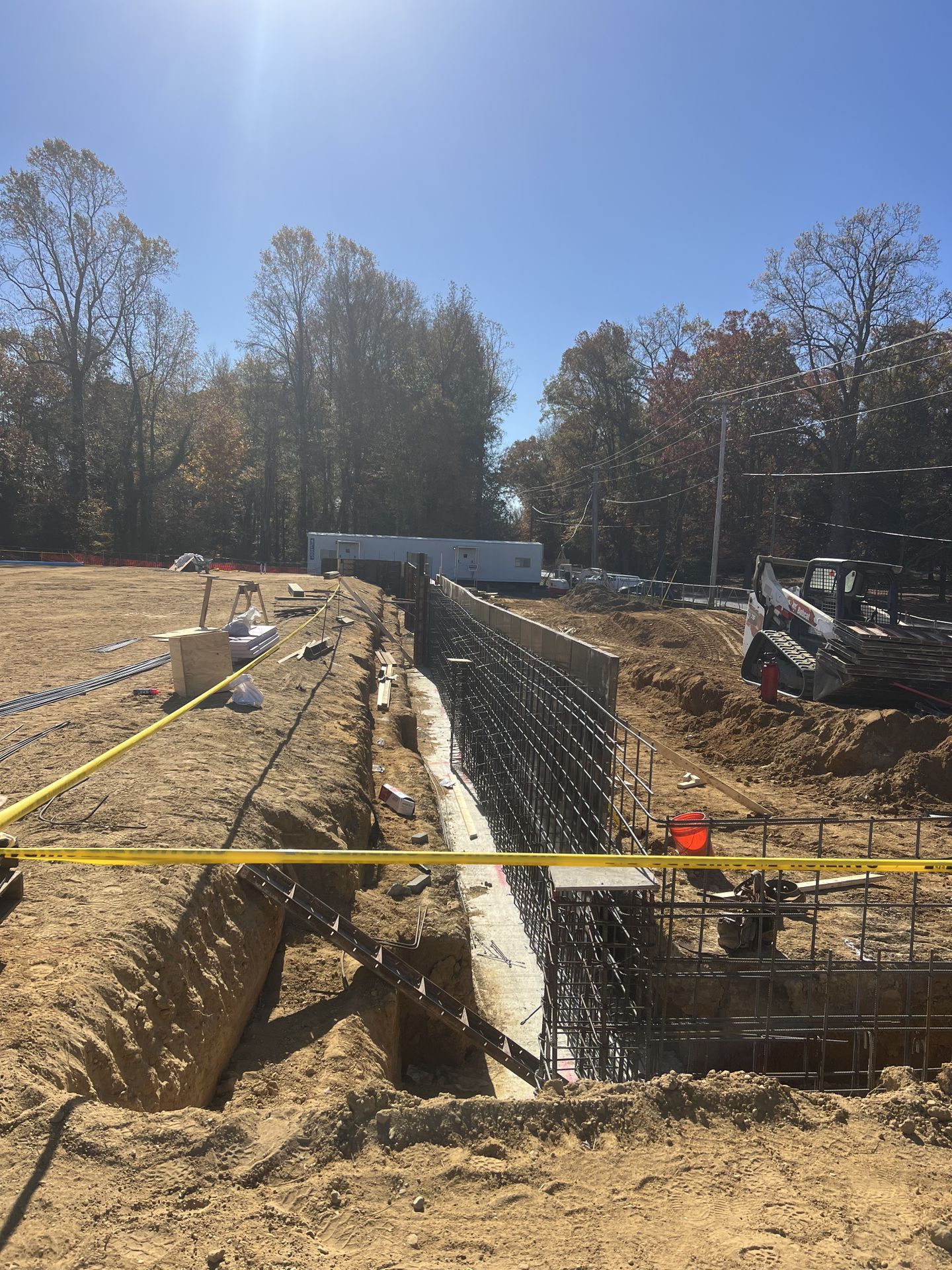 Construction site with a deep trench, rebar, caution tape, and an excavator beside a wooded area.