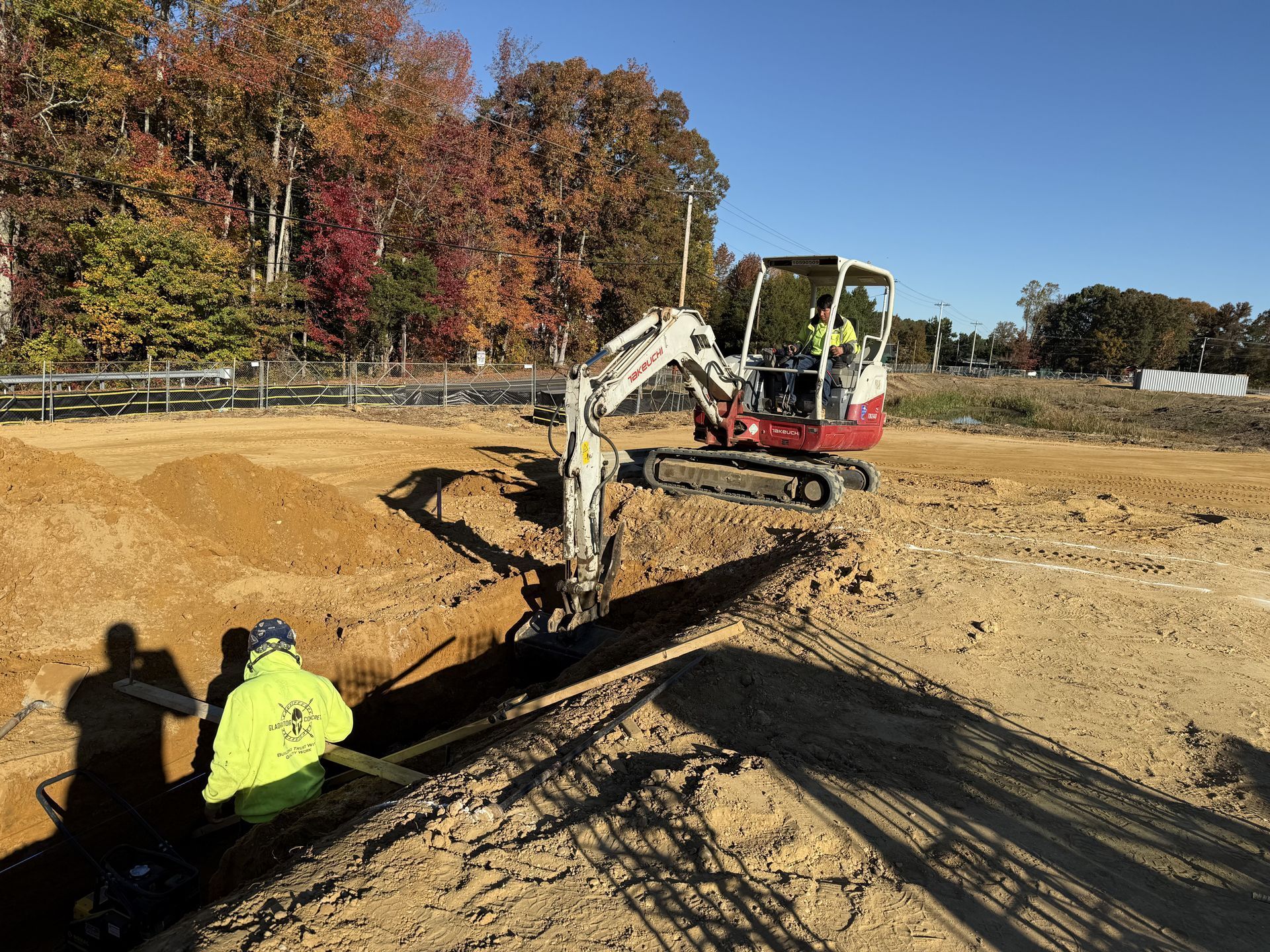 Excavator working in a dirt trench on a construction site, with a worker in a neon jacket nearby.