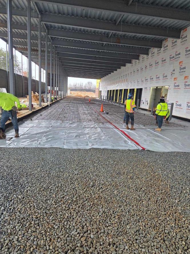 Workers spreading gravel inside a covered construction site under a metal roof