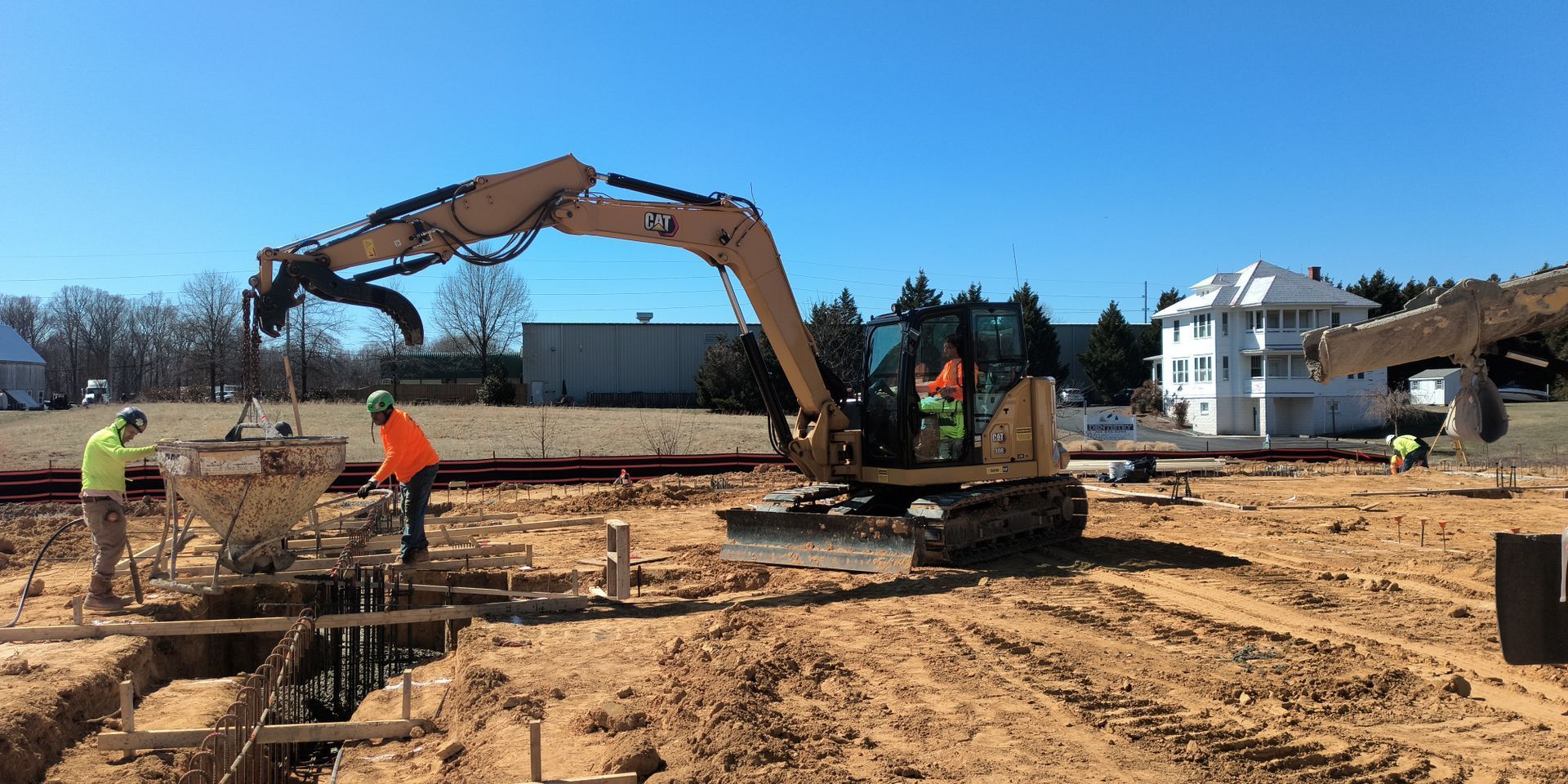 Excavator at a muddy construction site with workers and building foundations in the background