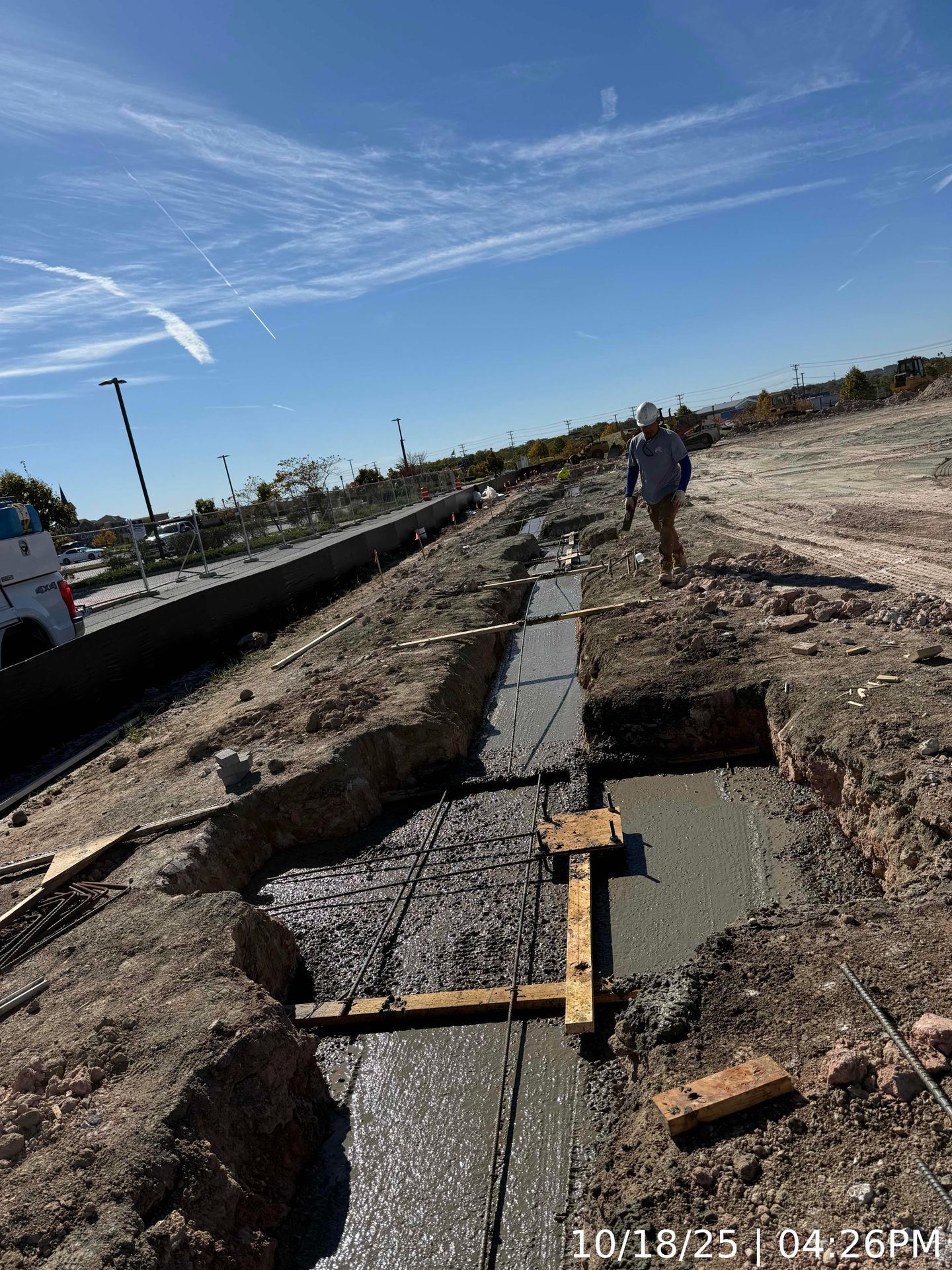 Roadside concrete ditch under construction, with worker and truck beside a dirt path under a clear blue sky.