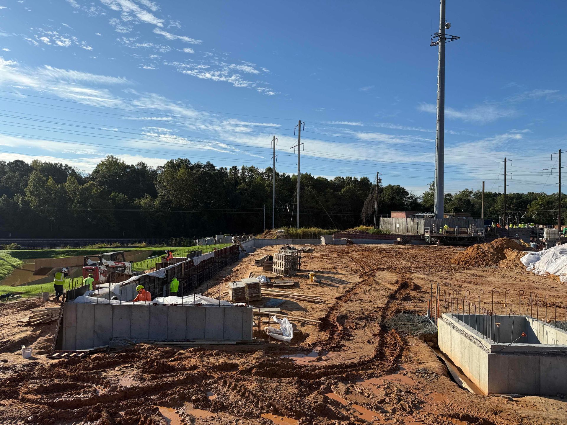 Construction site with dirt roads, utility poles, and concrete foundations under a partly cloudy sky