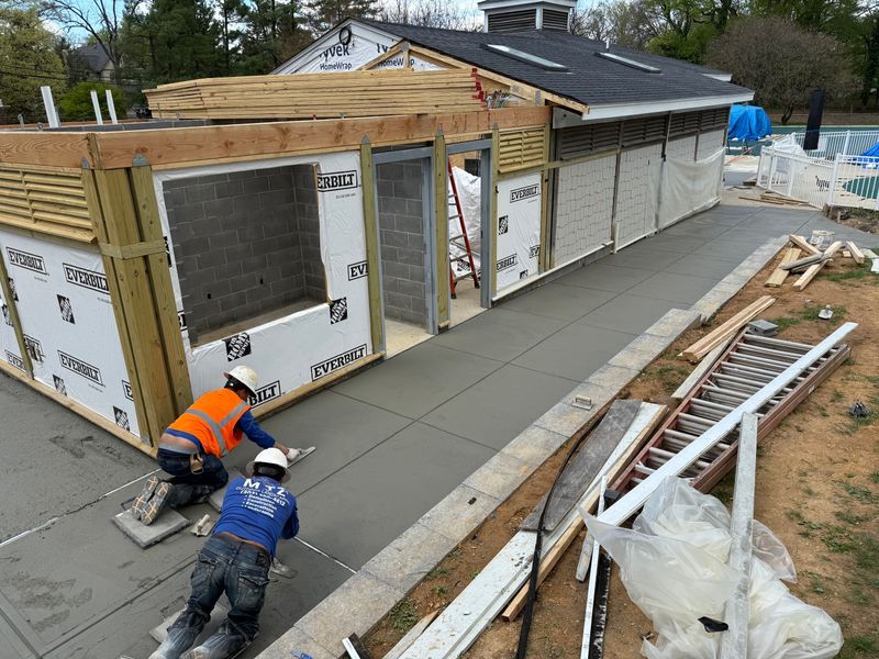 Workers installing insulation on a house under construction, with a wooden roof frame and paved walkway.