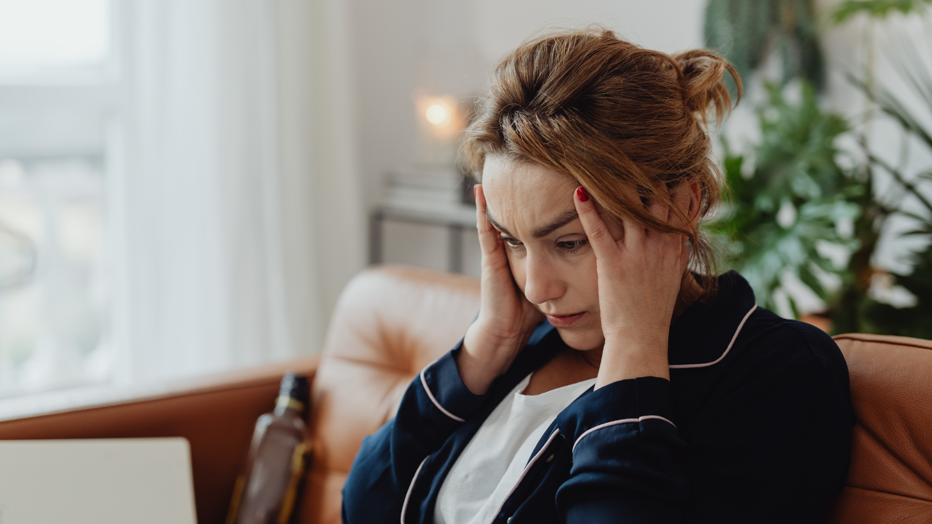 Woman in a dark jacket sits on a couch, holding her head and looking stressed in a bright room.