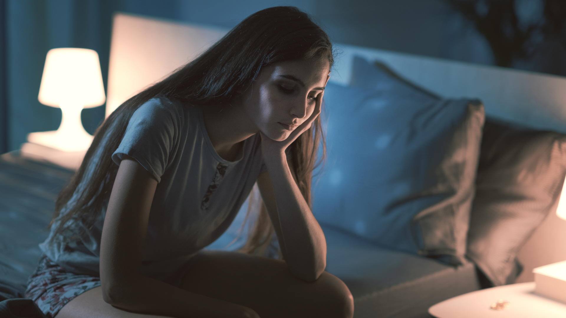 Woman sitting on a bed at night, looking pensive beside a glowing bedside lamp.