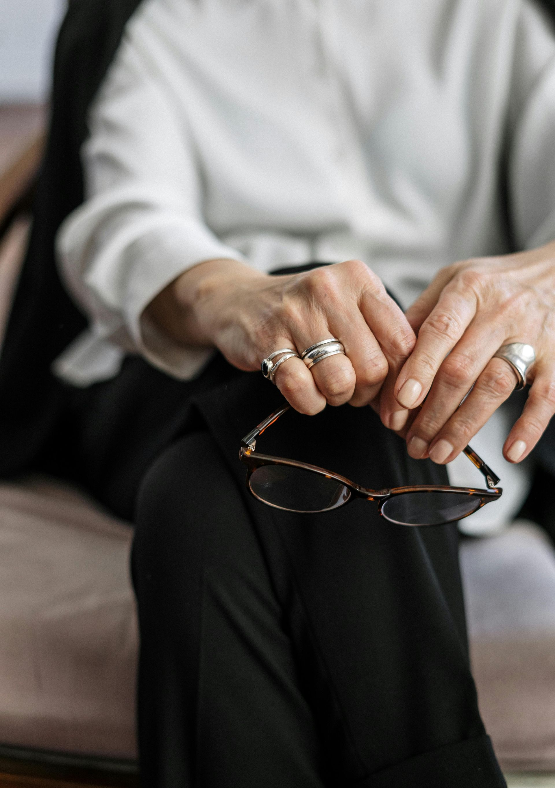 Person in white top holding eyeglasses over crossed black-clad legs on a sofa.