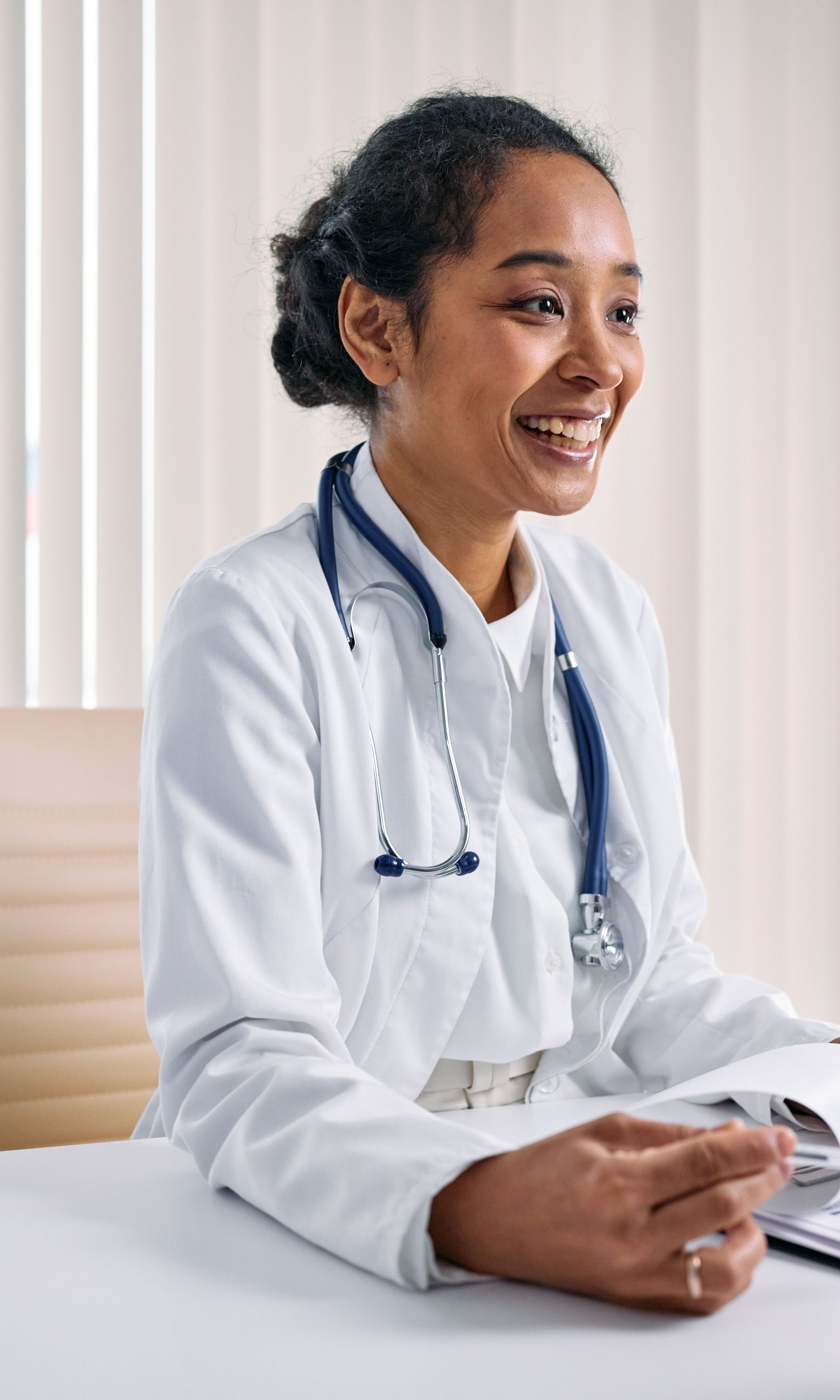 Smiling doctor in a white coat with a stethoscope, seated at a desk in a bright clinic