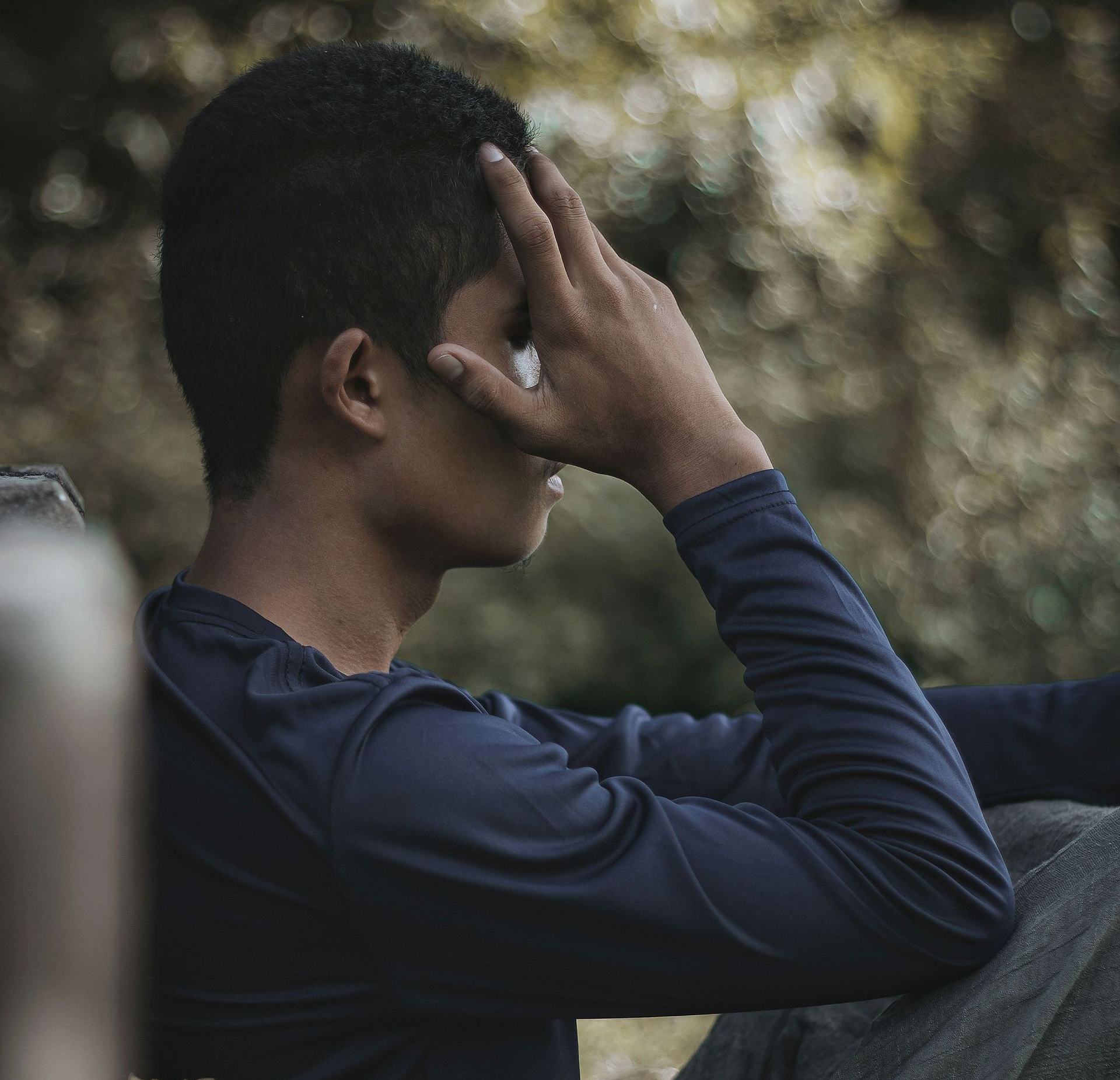 Person in a navy shirt sits outdoors with a hand covering their face in a contemplative pose