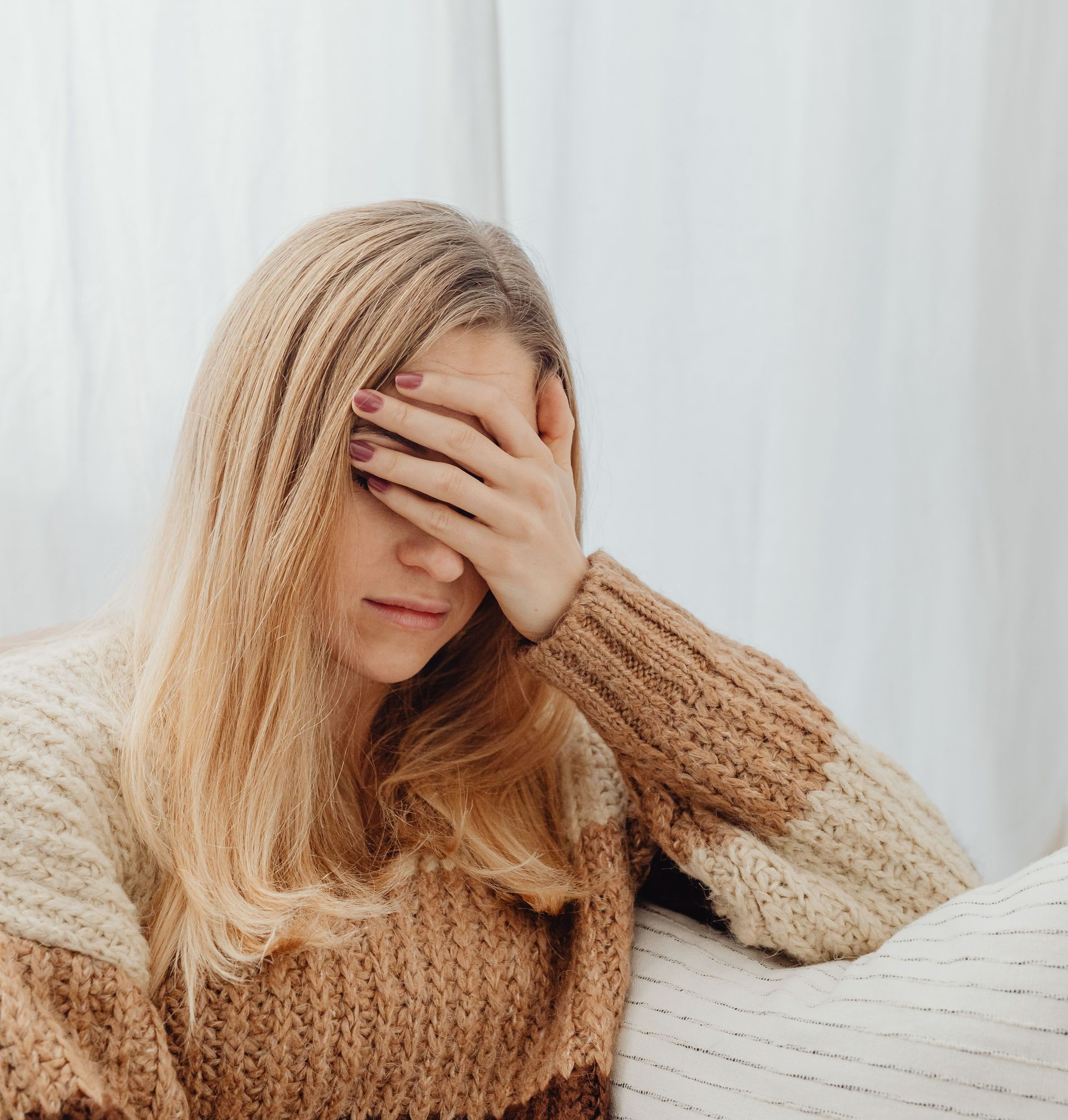 Woman in a beige sweater sitting on a couch, covering her face with one hand.