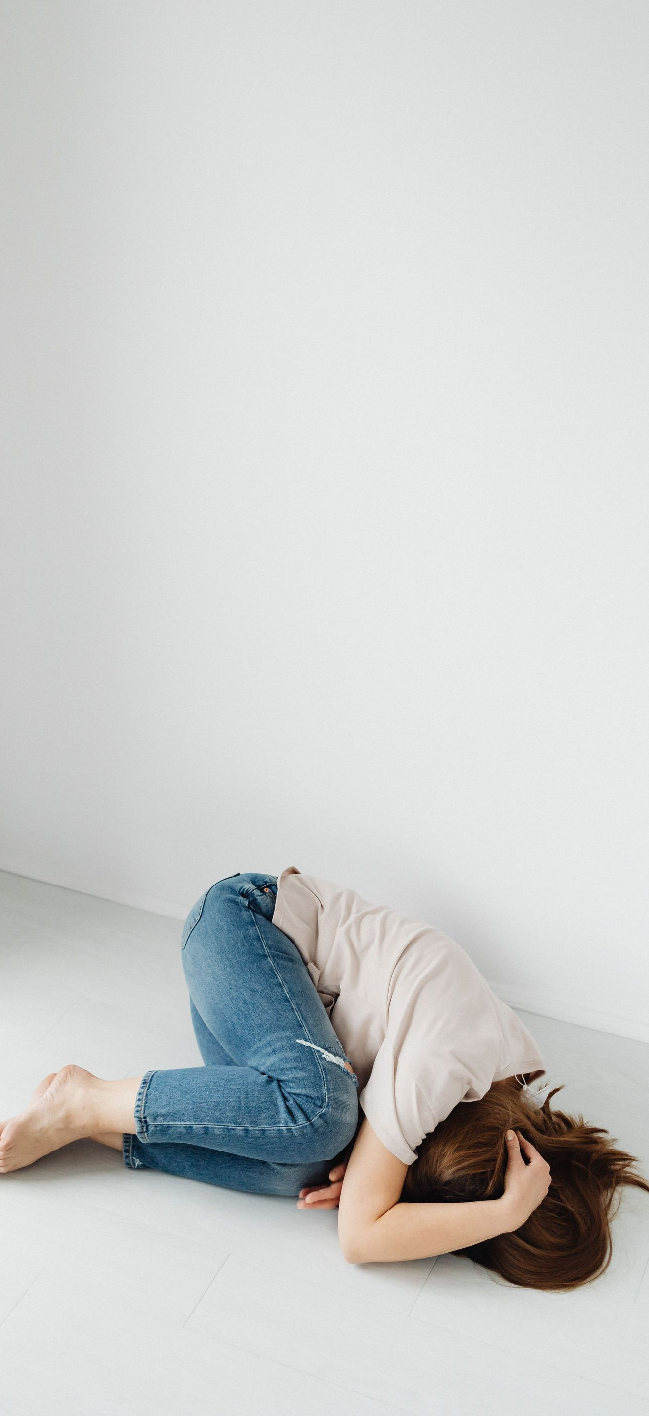 Person curled on white floor against wall, knees tucked in, covering face with hands.