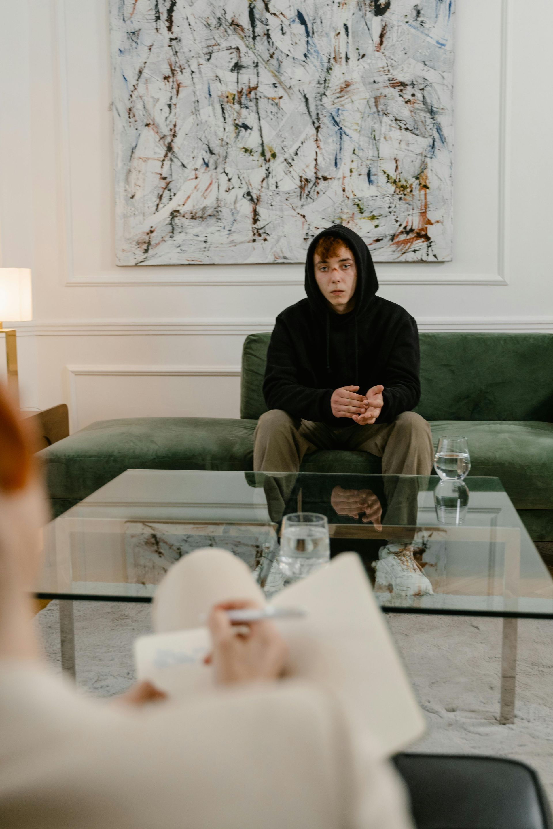 Person in a black hoodie sits on a green couch behind a glass coffee table in a bright living room.