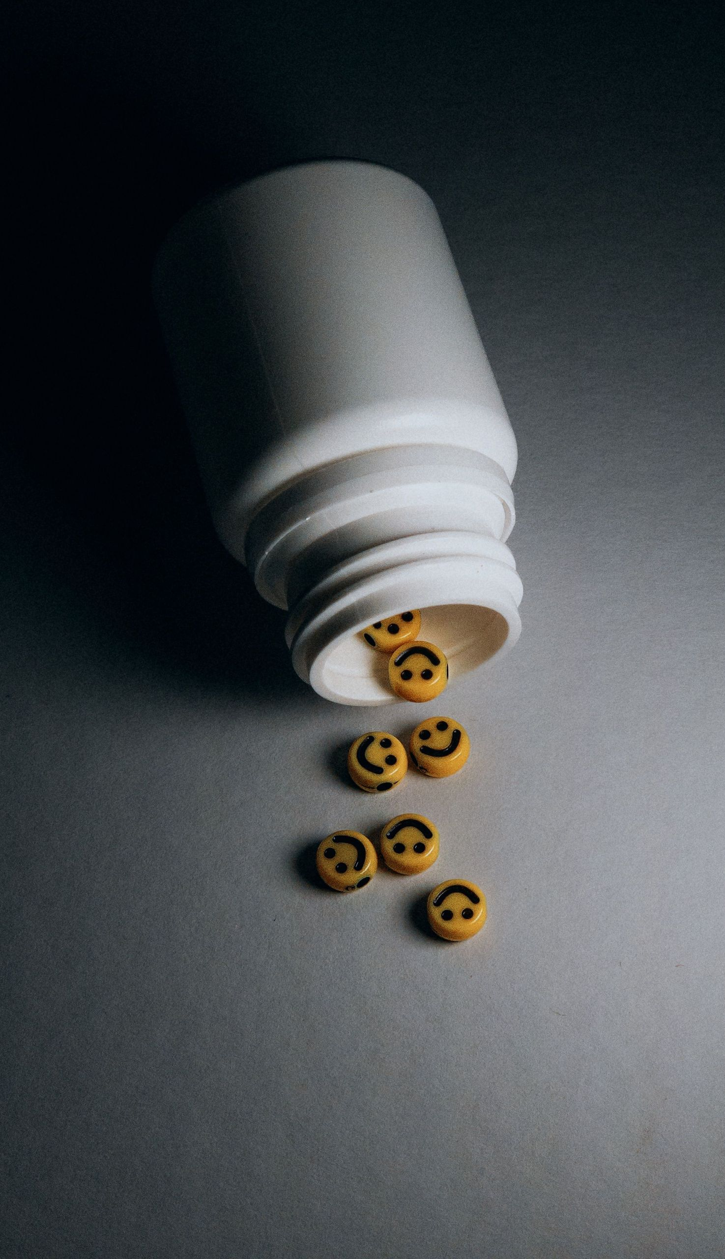White pill bottle tipped over, spilling small smiley-face tablets onto a gray surface.