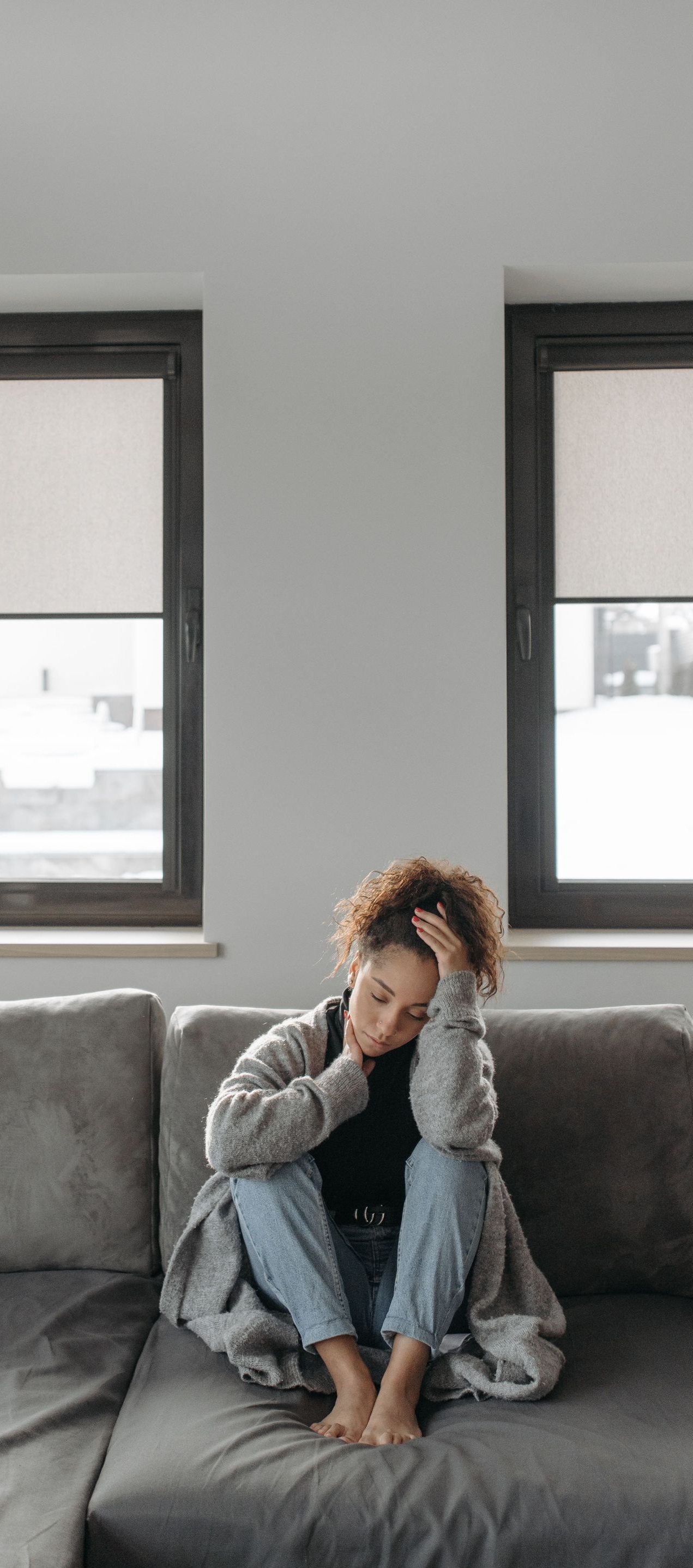 Person sitting curled up on a gray couch beneath two windows in a bright living room