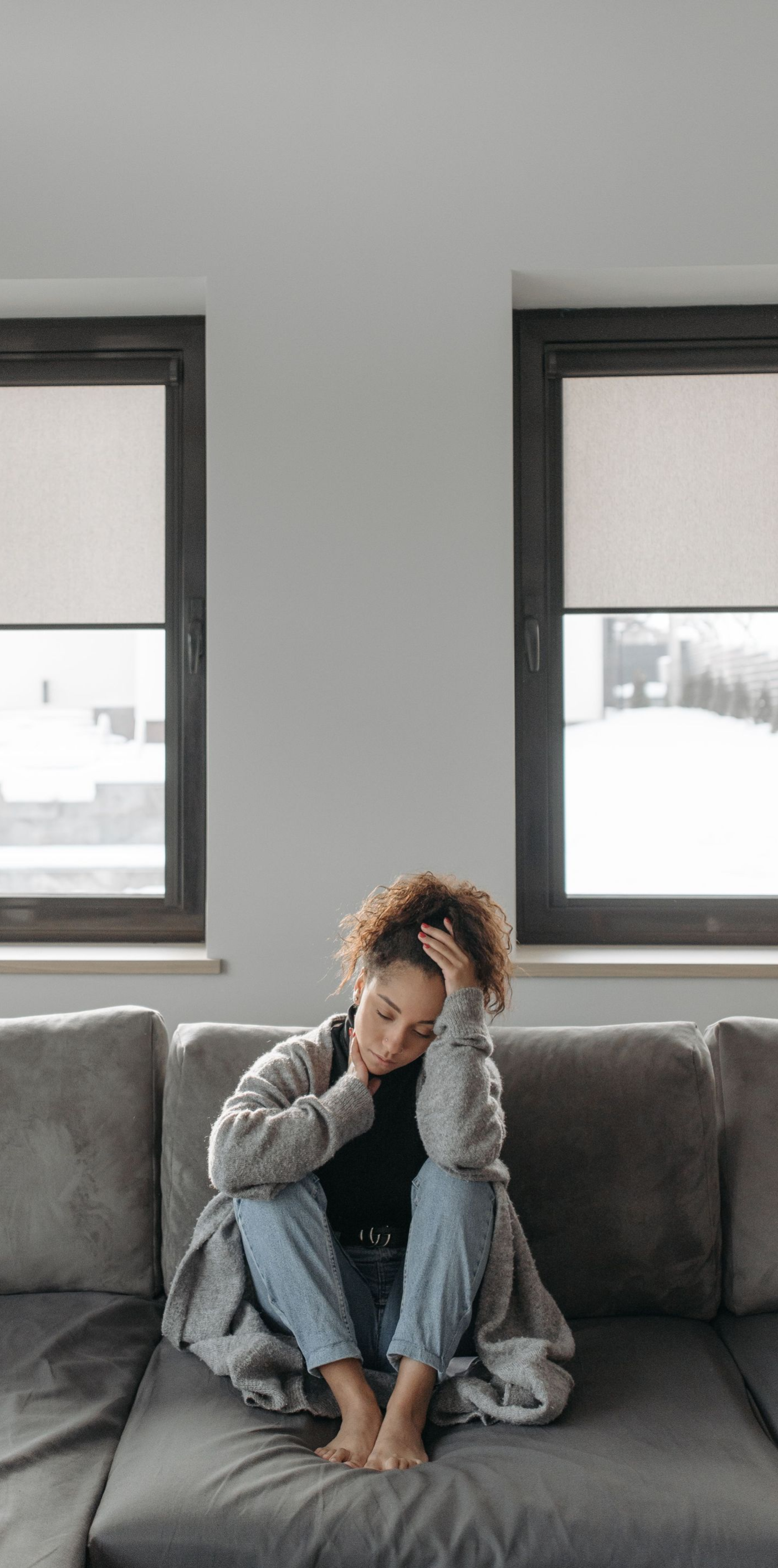 Person sitting curled up on a gray couch between two windows, holding their head in a dim living room